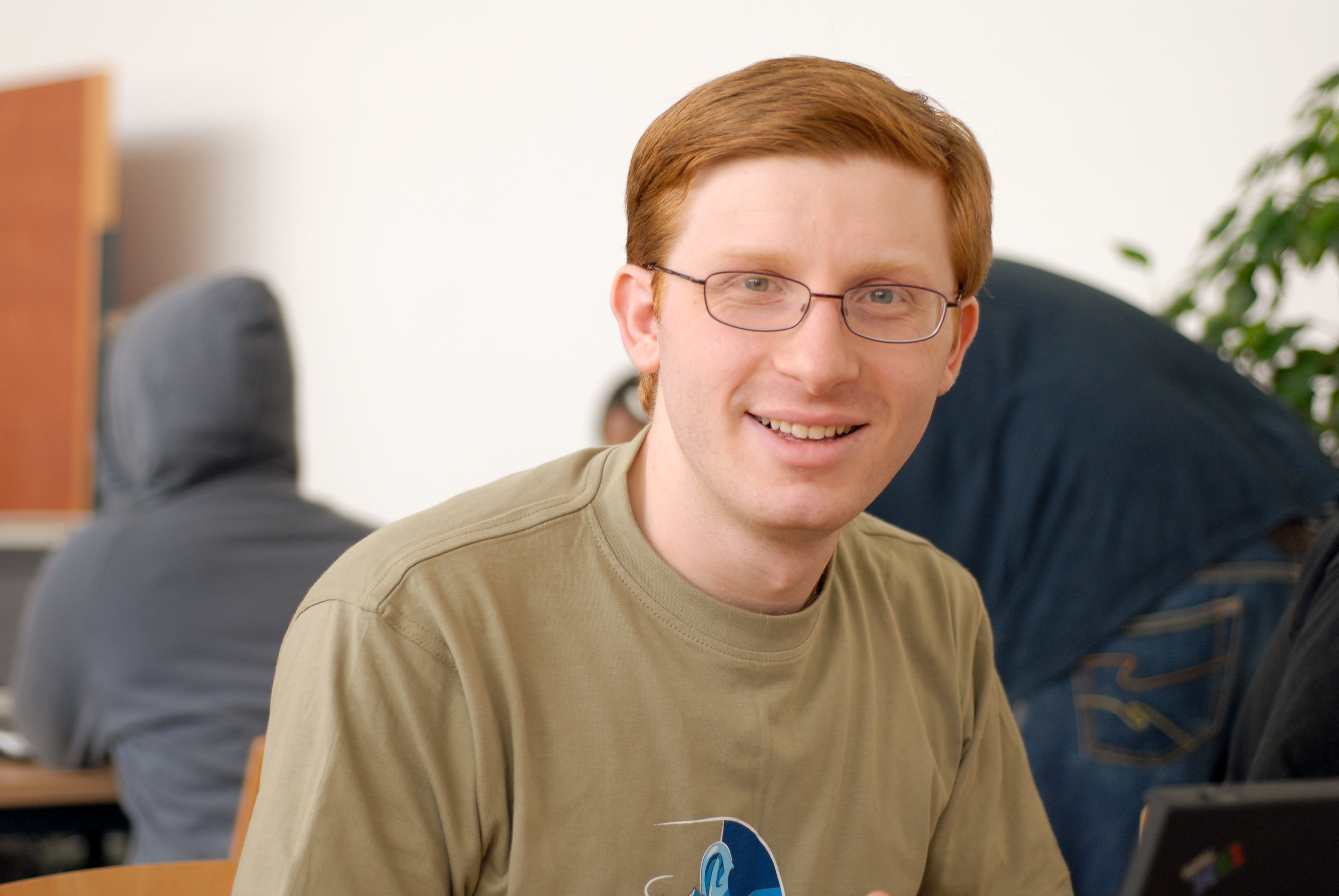 A man with glasses and a green t-shirt smiles at the camera while sitting in a busy indoor setting.