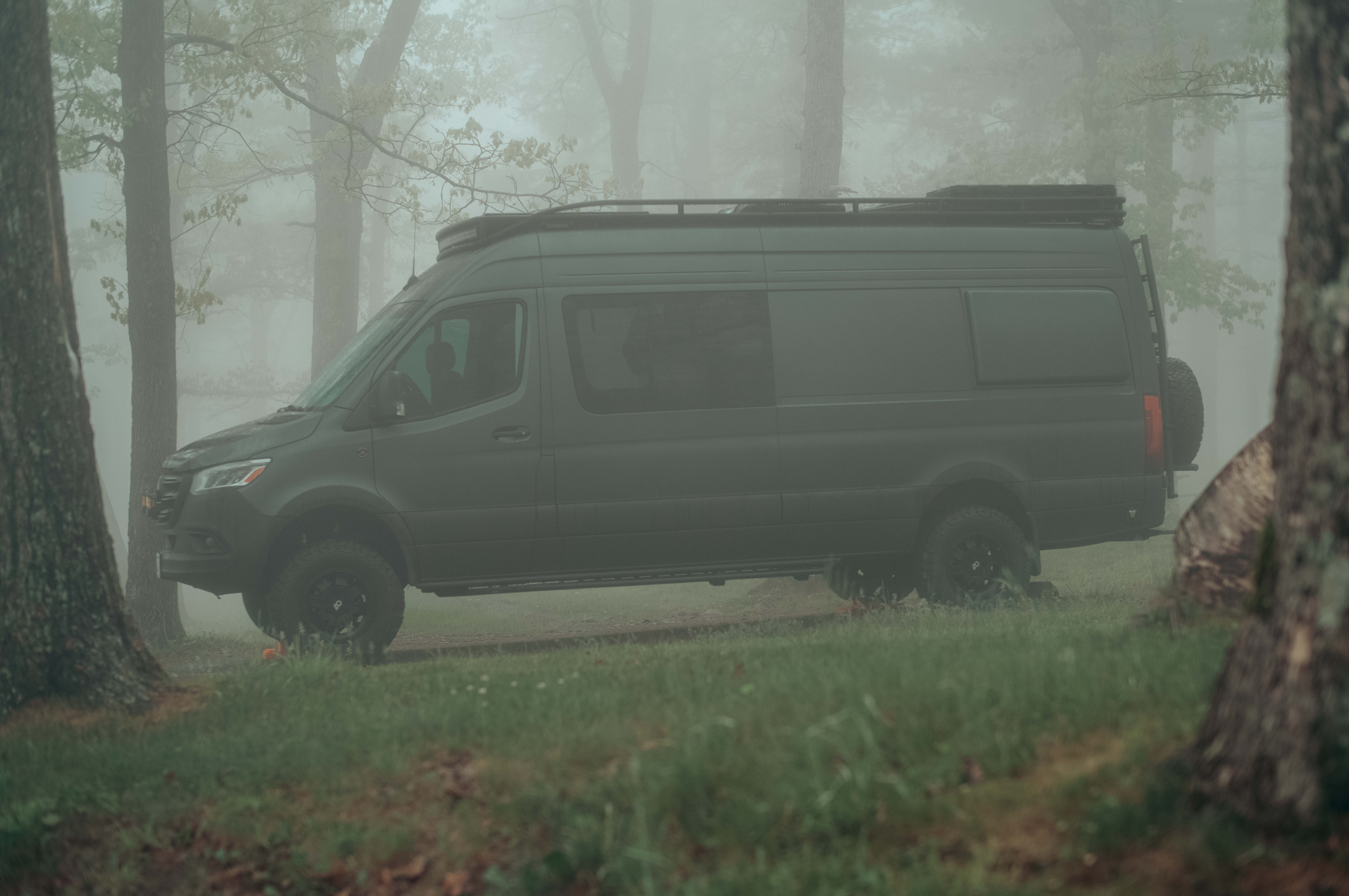 Matte black camper van parked among trees in dense morning fog.