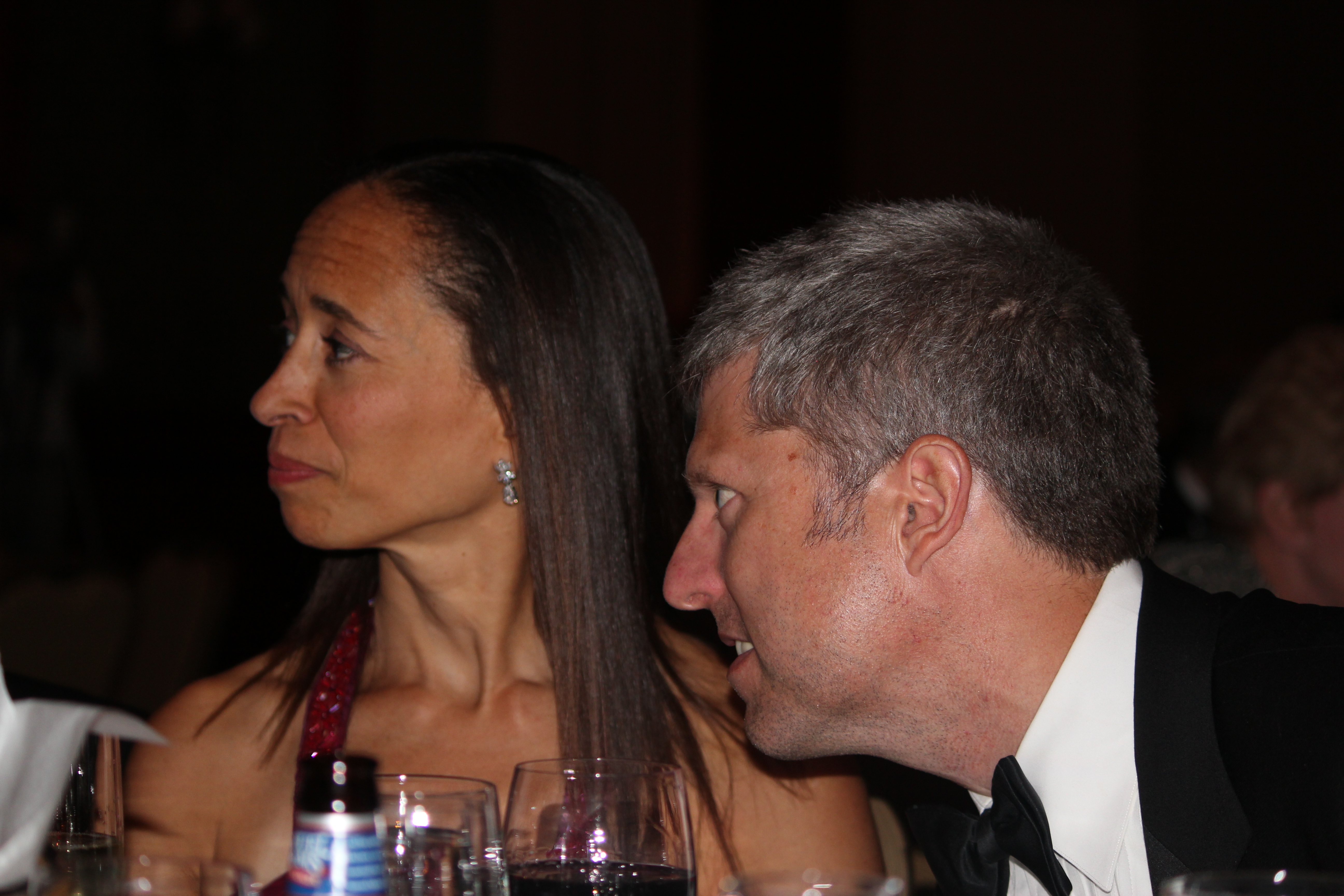 A man in a tuxedo leans in to speak to a woman in a red dress at a banquet.