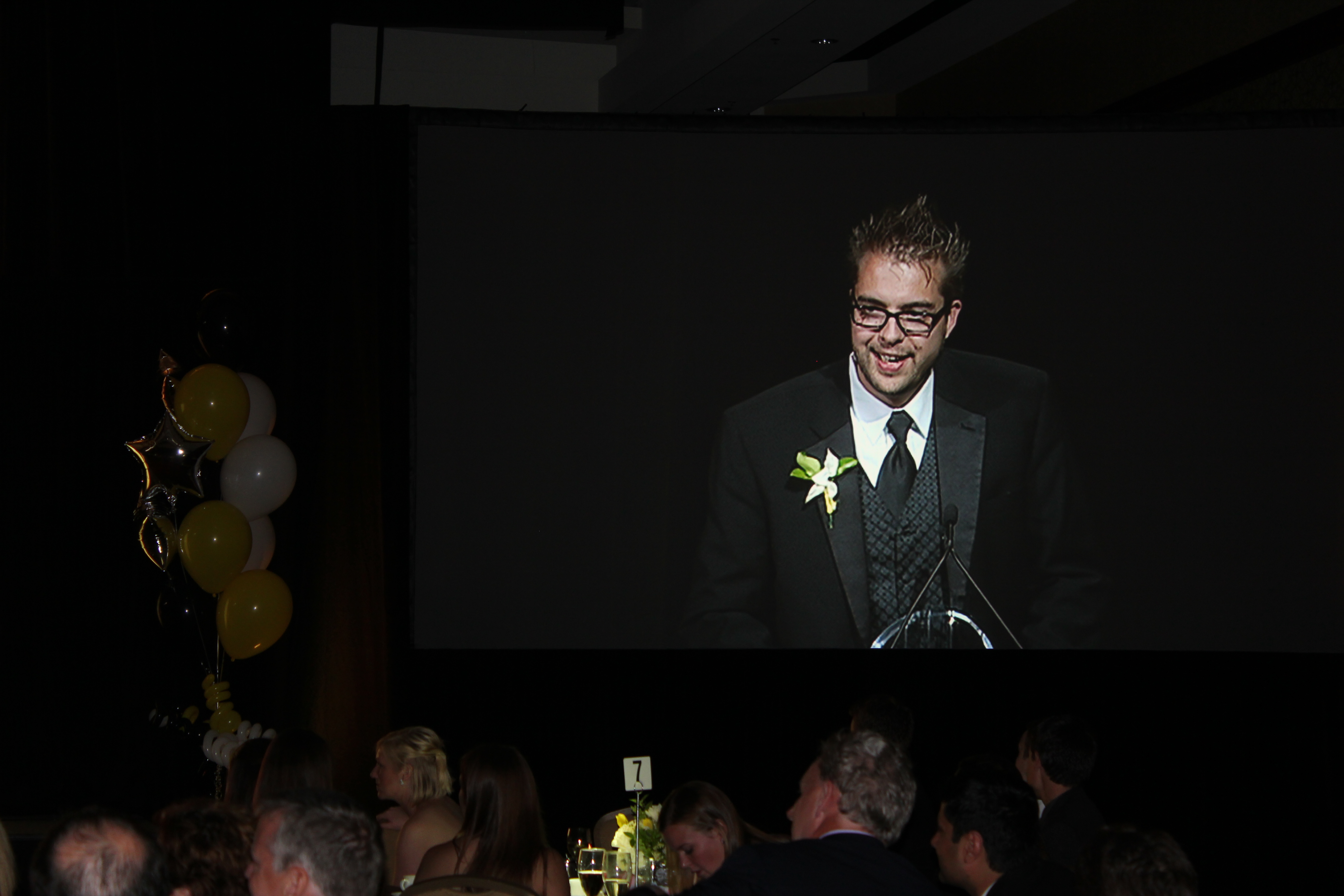A speaker in a tuxedo stands at a podium, addressing the audience at an award banquet.