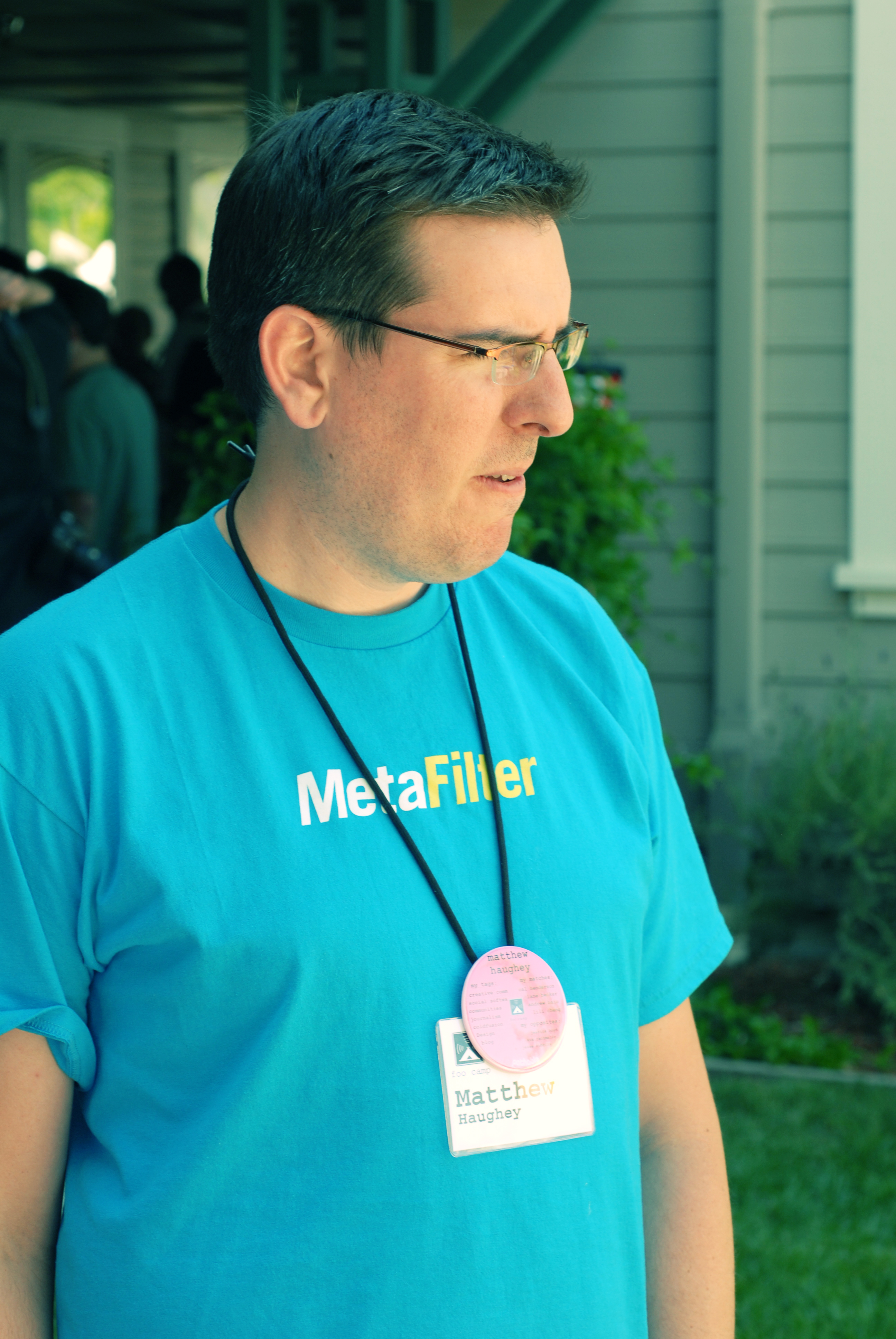 A man wearing a blue &quot;MetaFilter&quot; shirt and name badge stands outside, looking to the side.