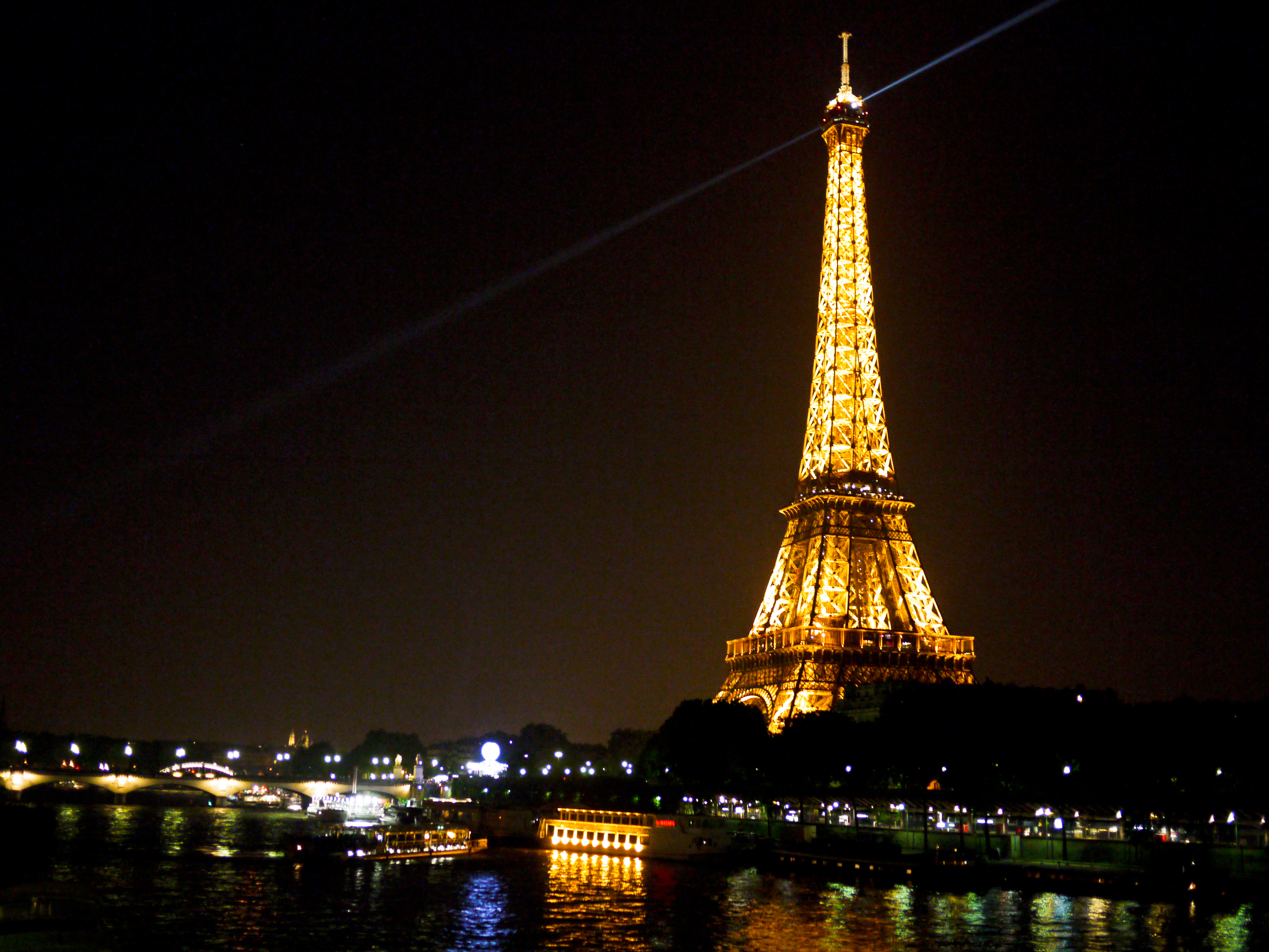 The Eiffel Tower glows at night with golden lights, a spotlight shining from the top, and boats on the river.
