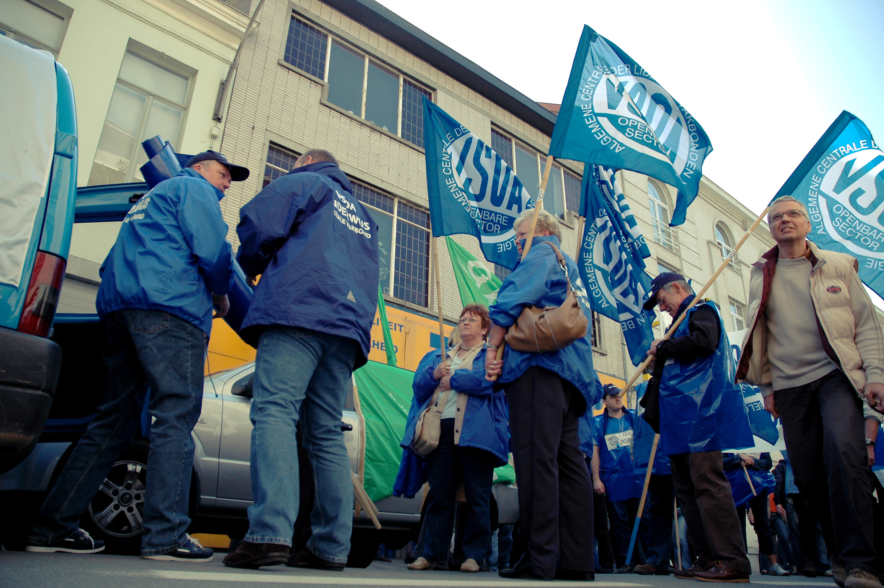 Protesters in blue jackets hold flags and gather on a city street during a demonstration.