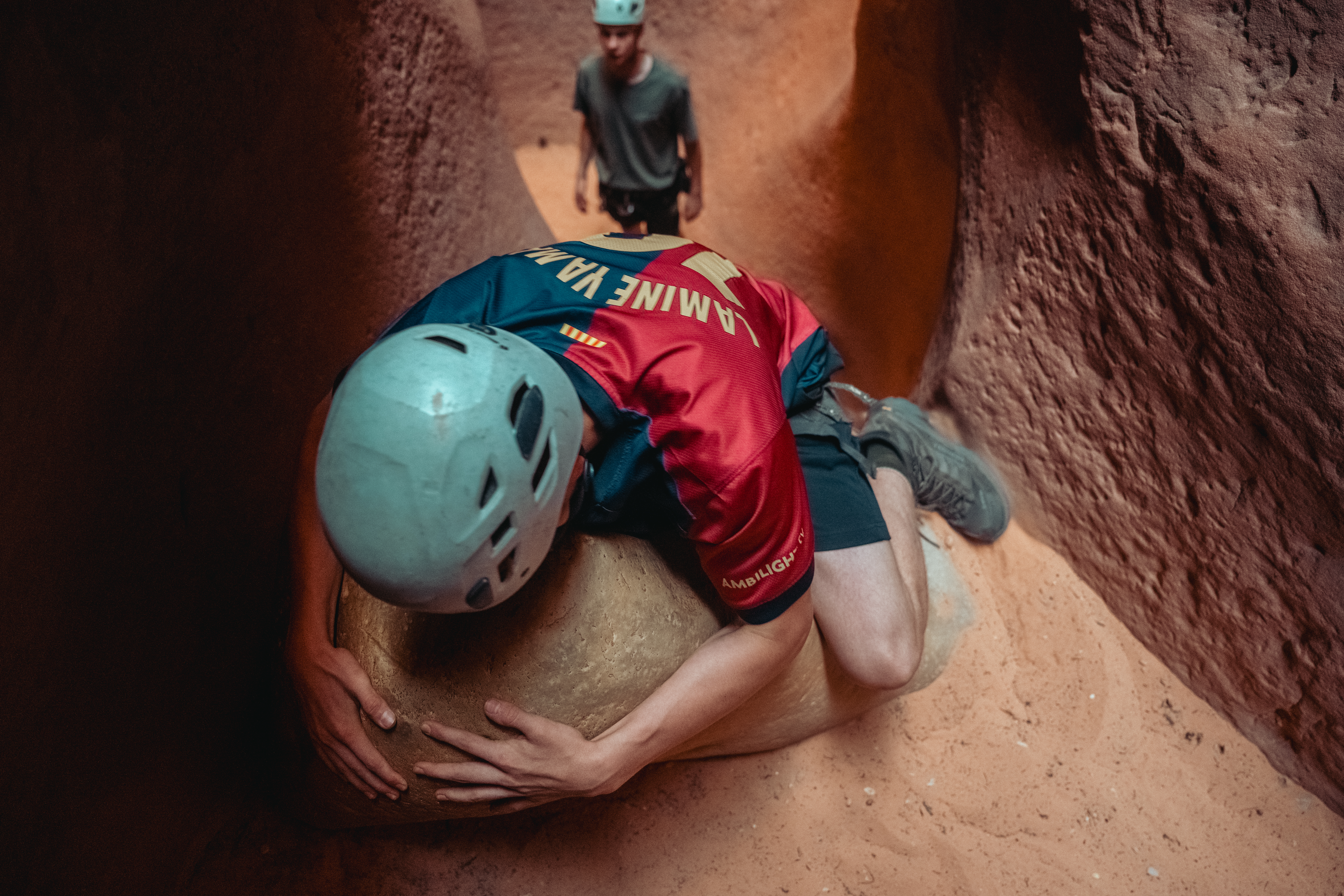 A teenager wearing a helmet lowering himself down while holding onto a large boulder inside a canyon in Zion National Park.