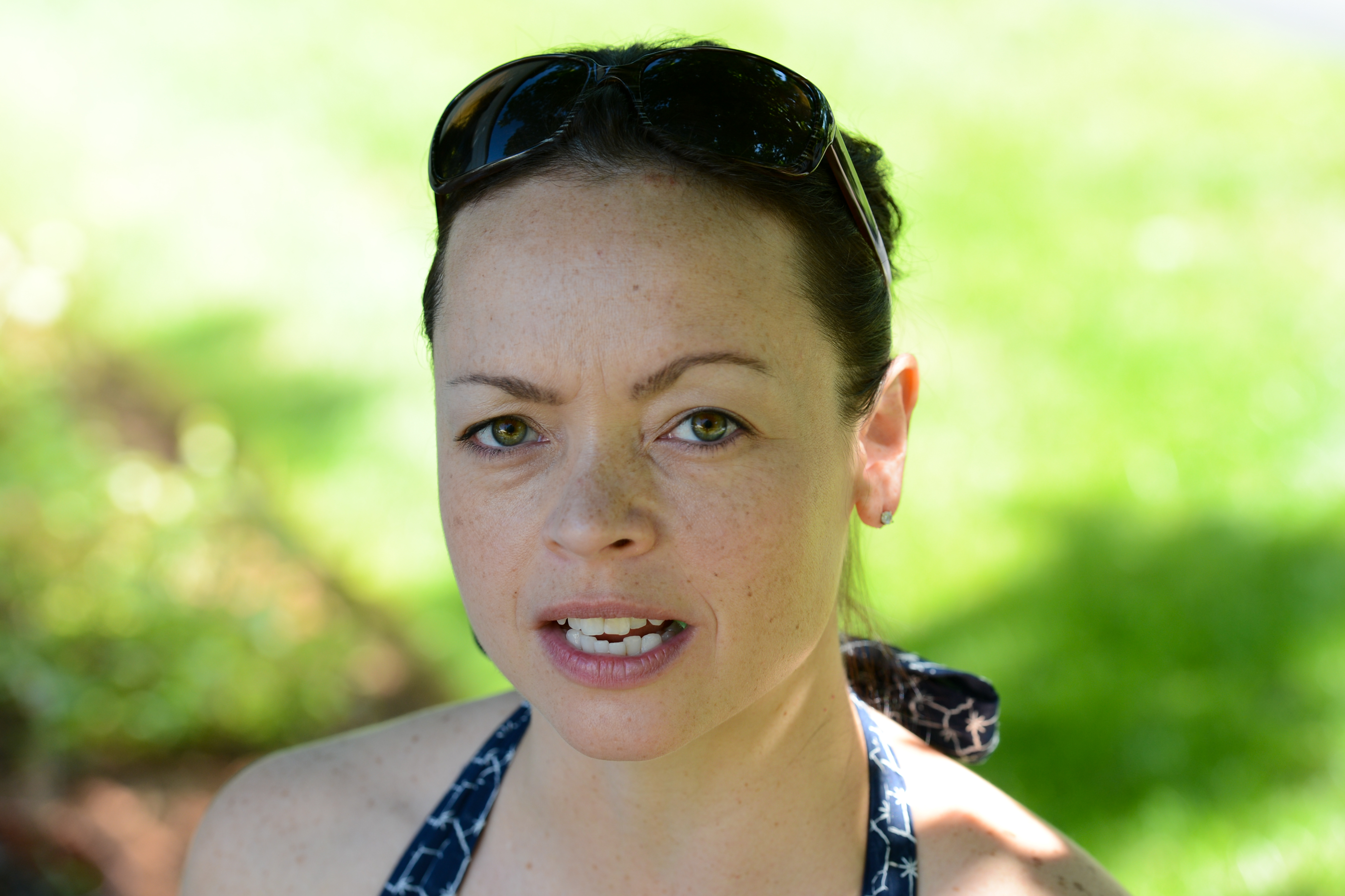 A woman with sunglasses on her head speaks while outdoors in a grassy area.