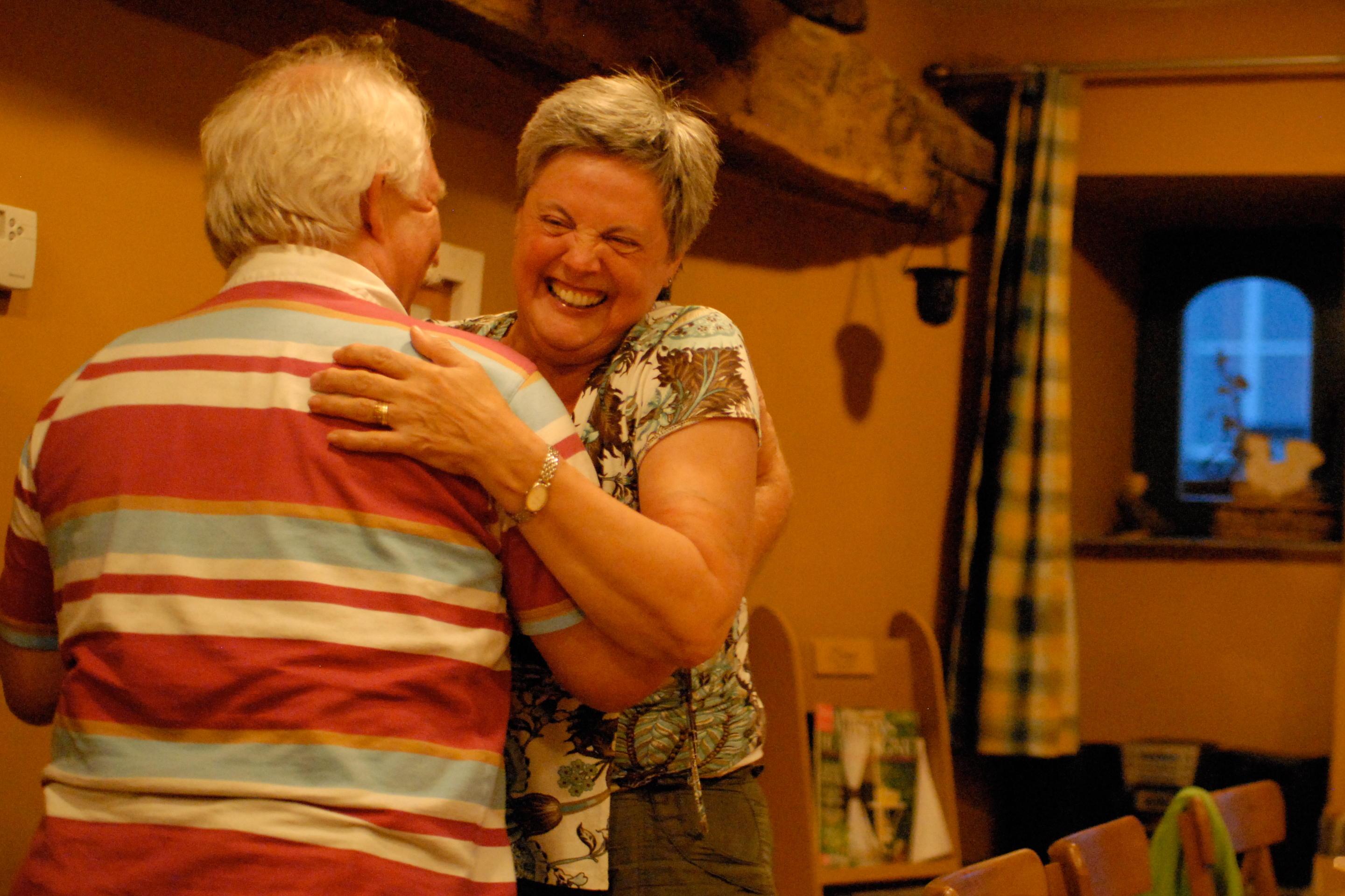 An elderly couple shares a joyful hug indoors, with the woman laughing and smiling warmly.