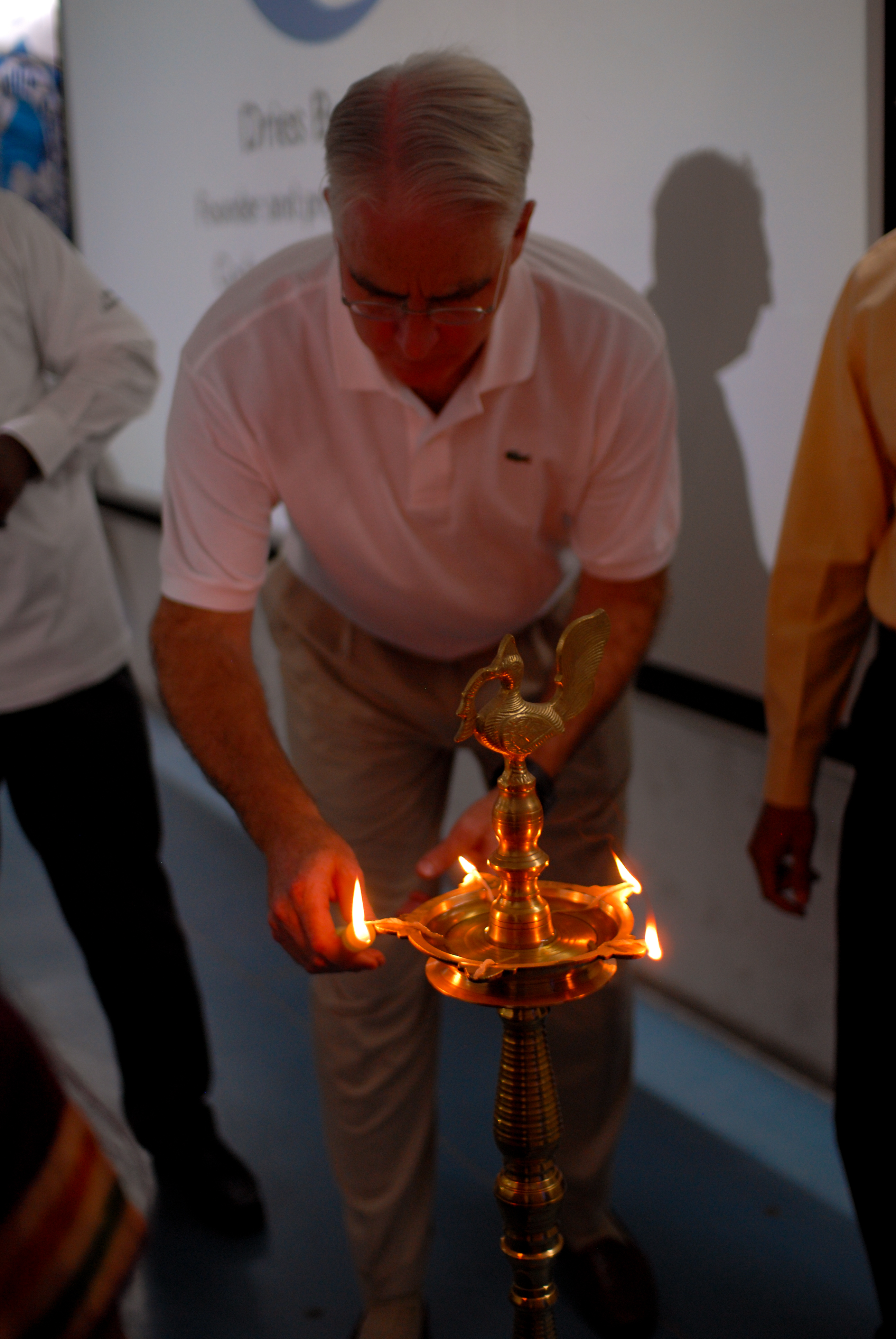 A man in a white shirt lights a traditional brass lamp at an event.