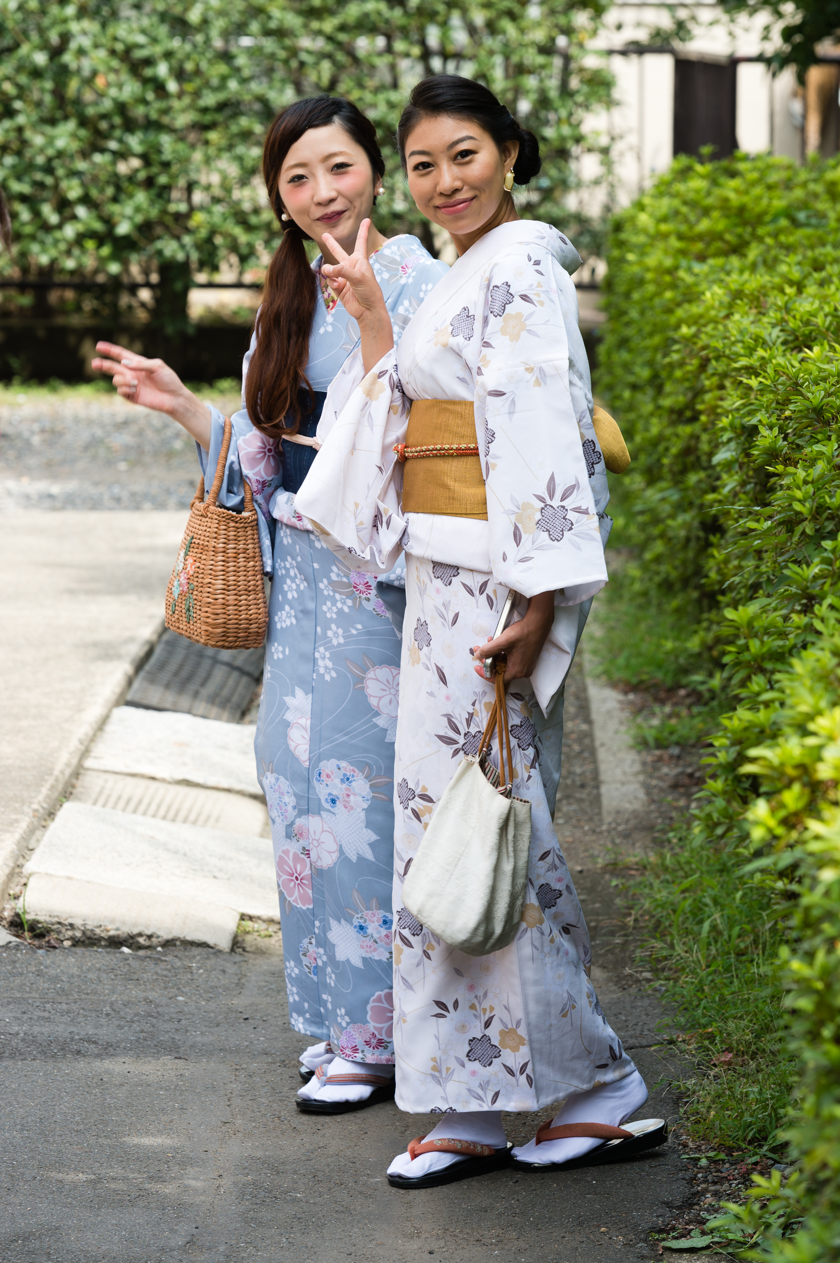 Two women wearing floral kimonos stand on a path, smiling and making peace signs while carrying handbags.
