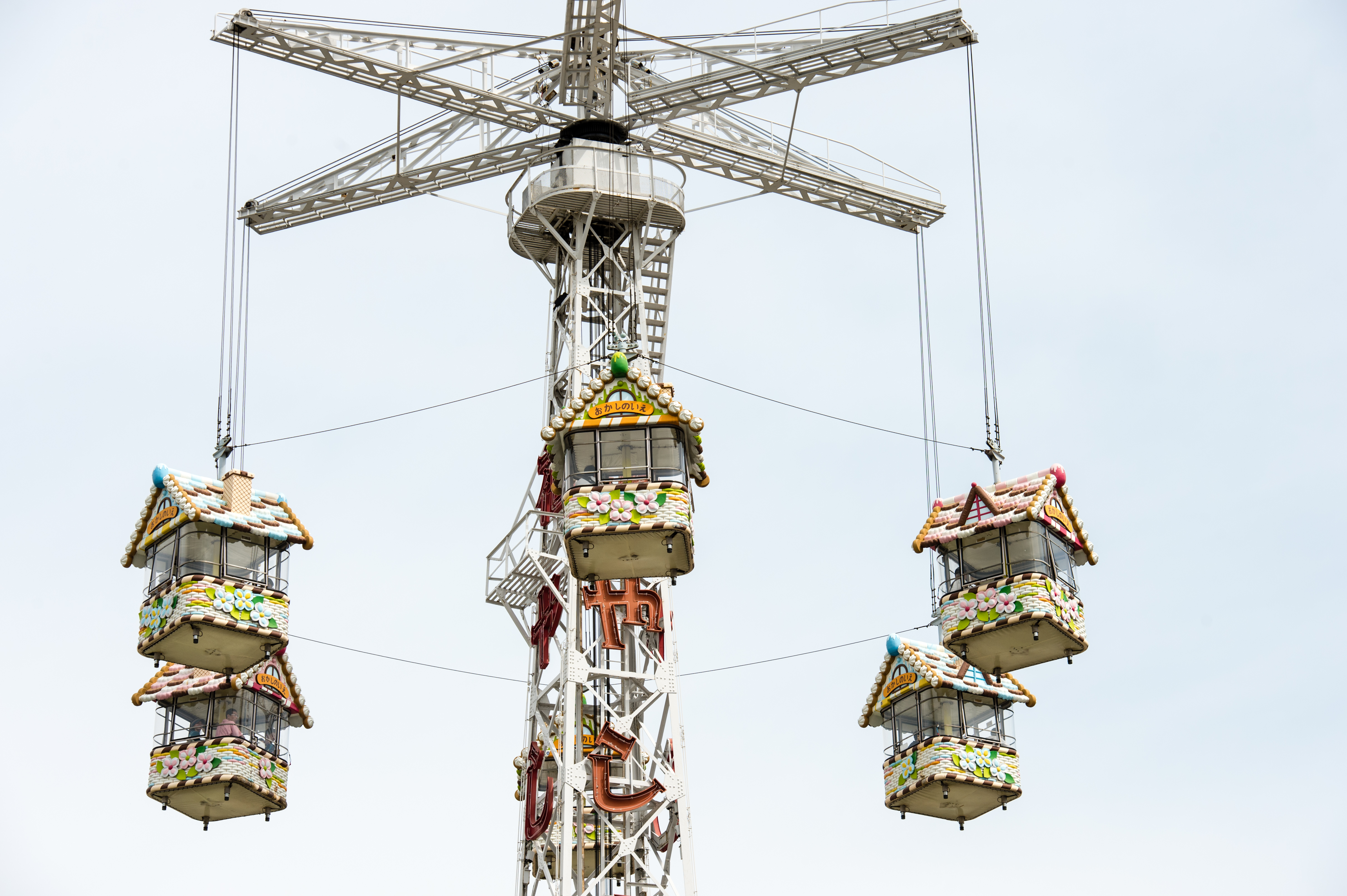 A tall amusement park ride with hanging gondolas shaped like colorful gingerbread houses, suspended by cables.