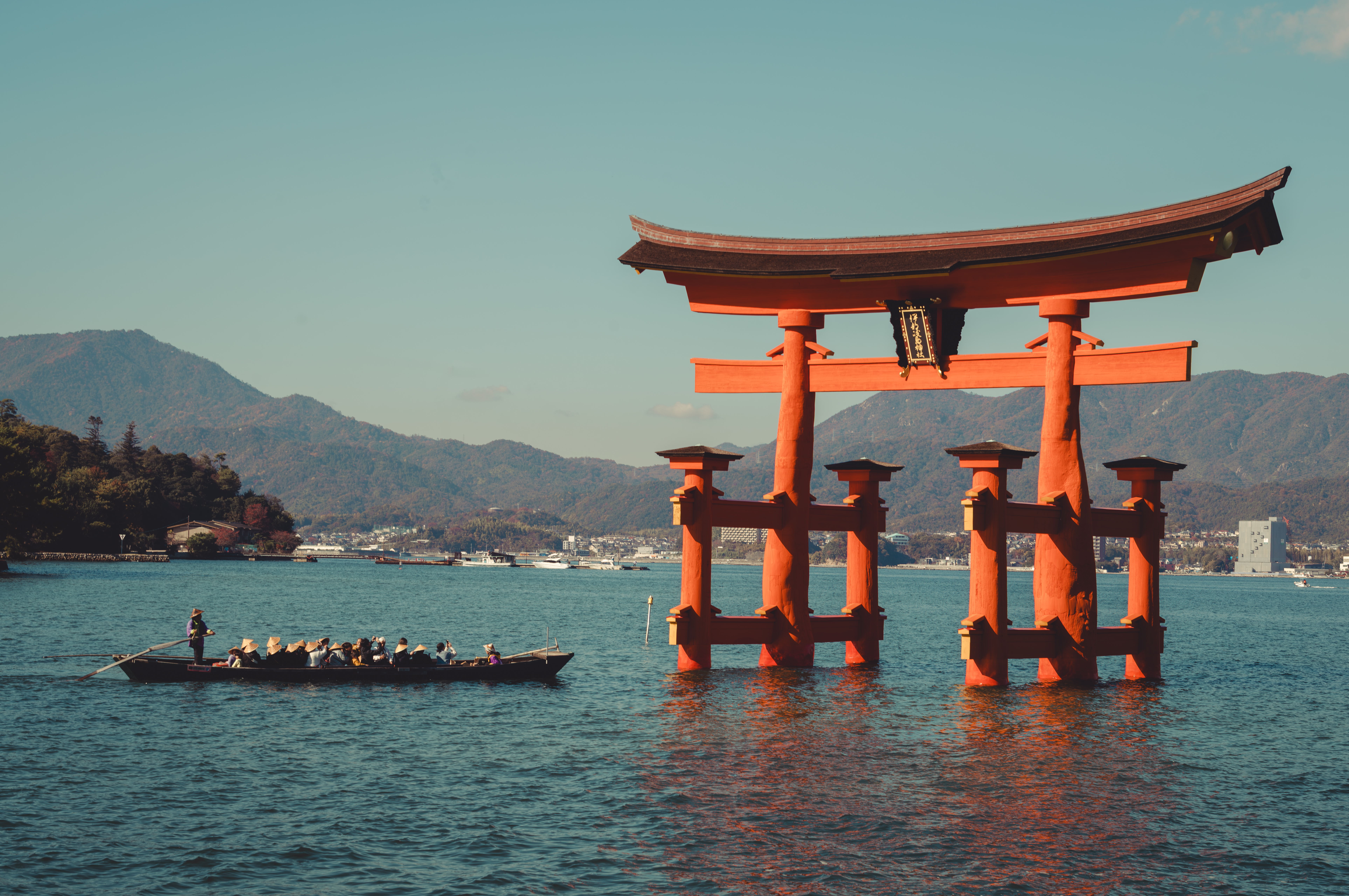 Large red torii gate stands in the water near a boat carrying people, with mountains and buildings in the background.