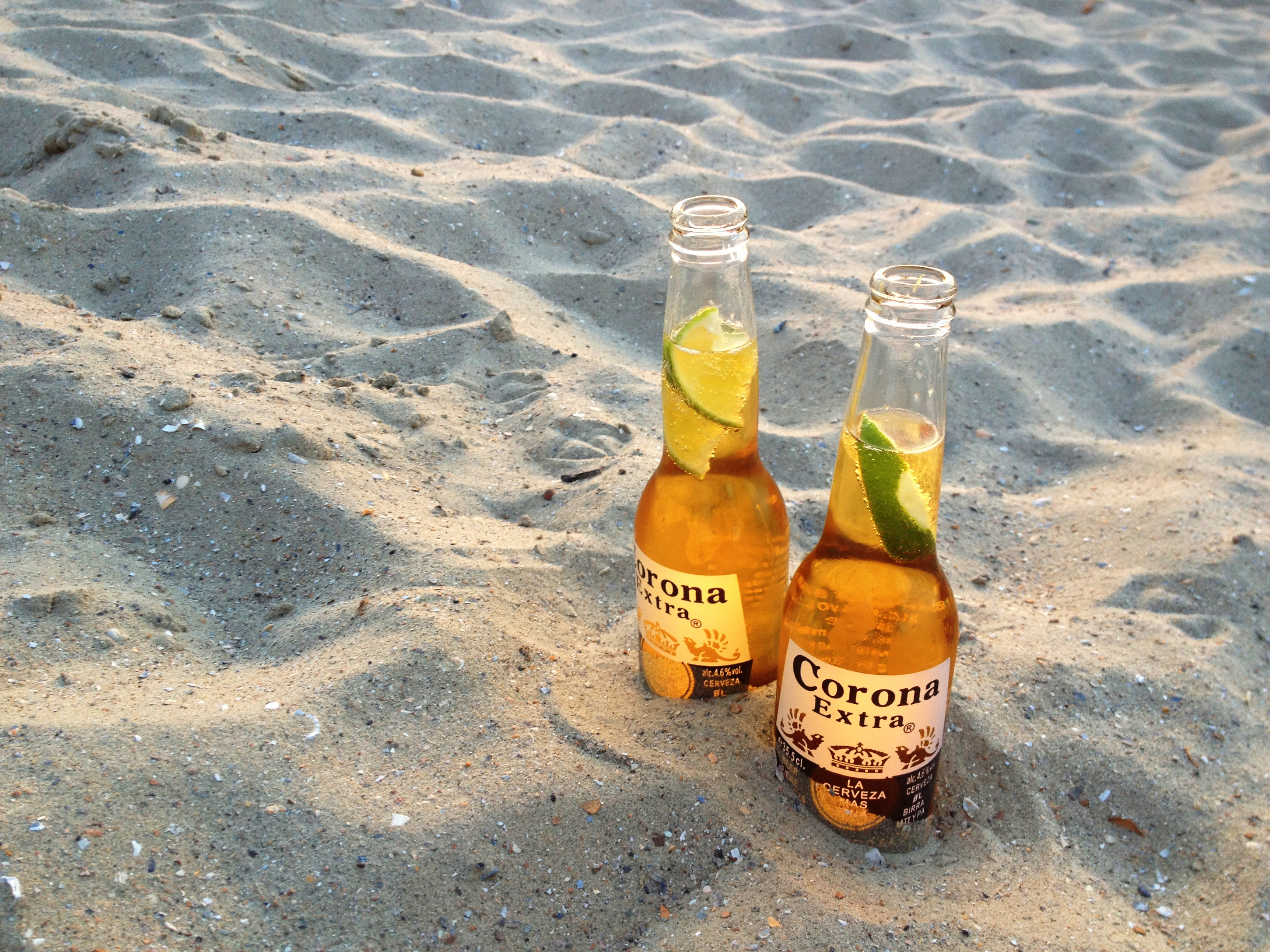 Two bottles of Corona beer with lime wedges sit on the sandy beach.