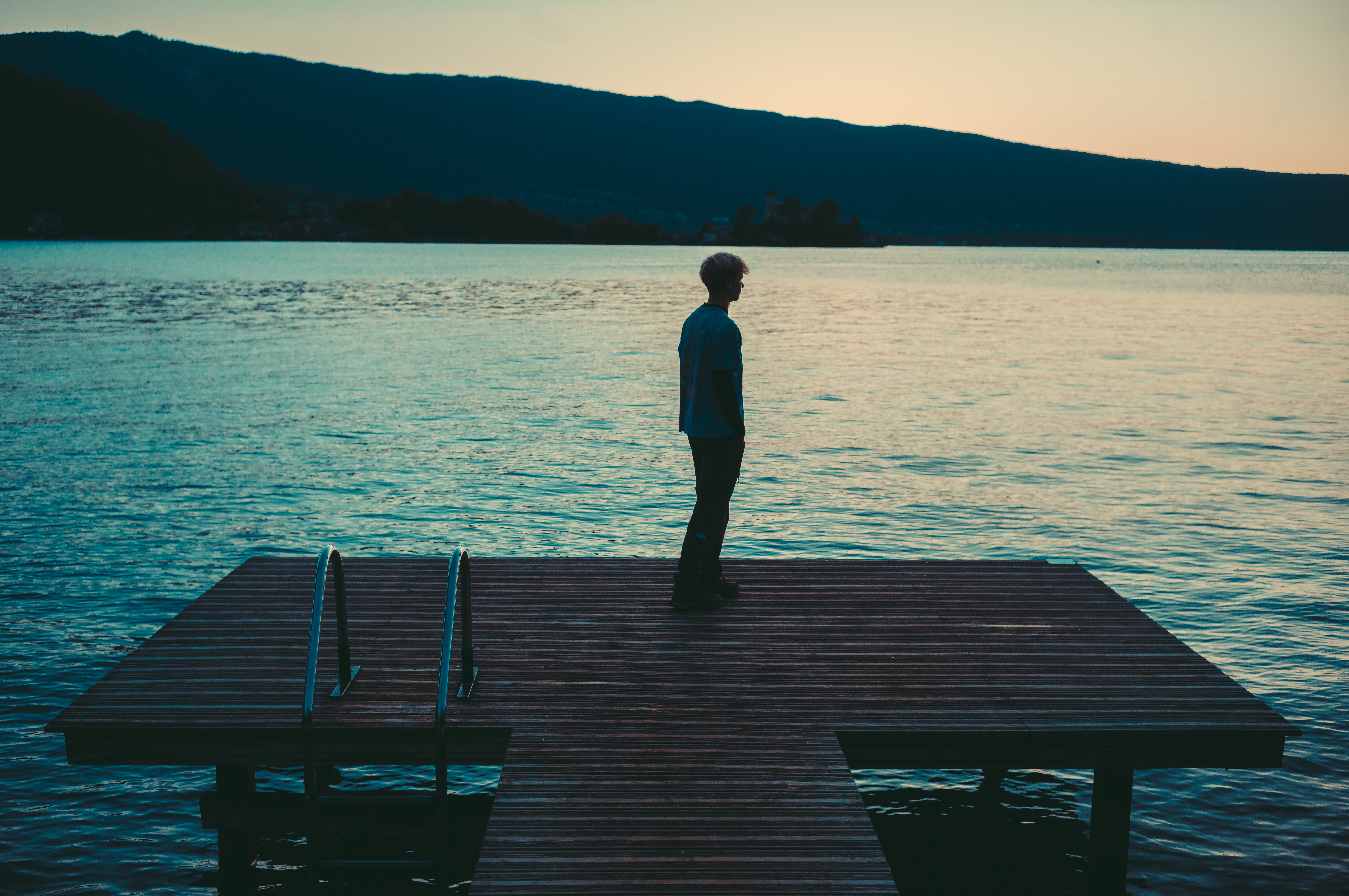 Person stands on a wooden dock at Lake Annecy, facing the water at dusk, with mountains in the background.