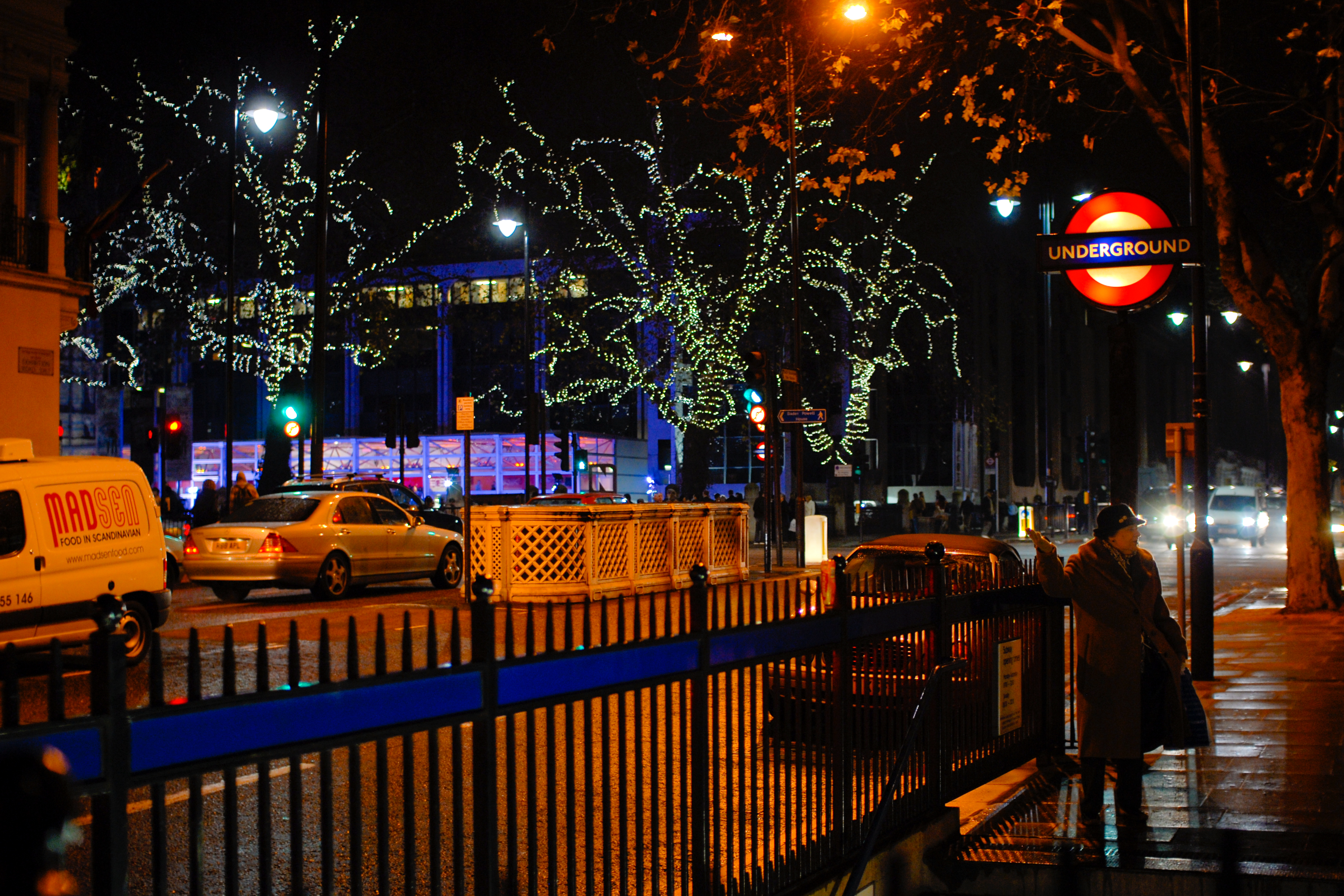 A person stands near a London Underground entrance at night, hailing a taxi on a busy, well-lit street.