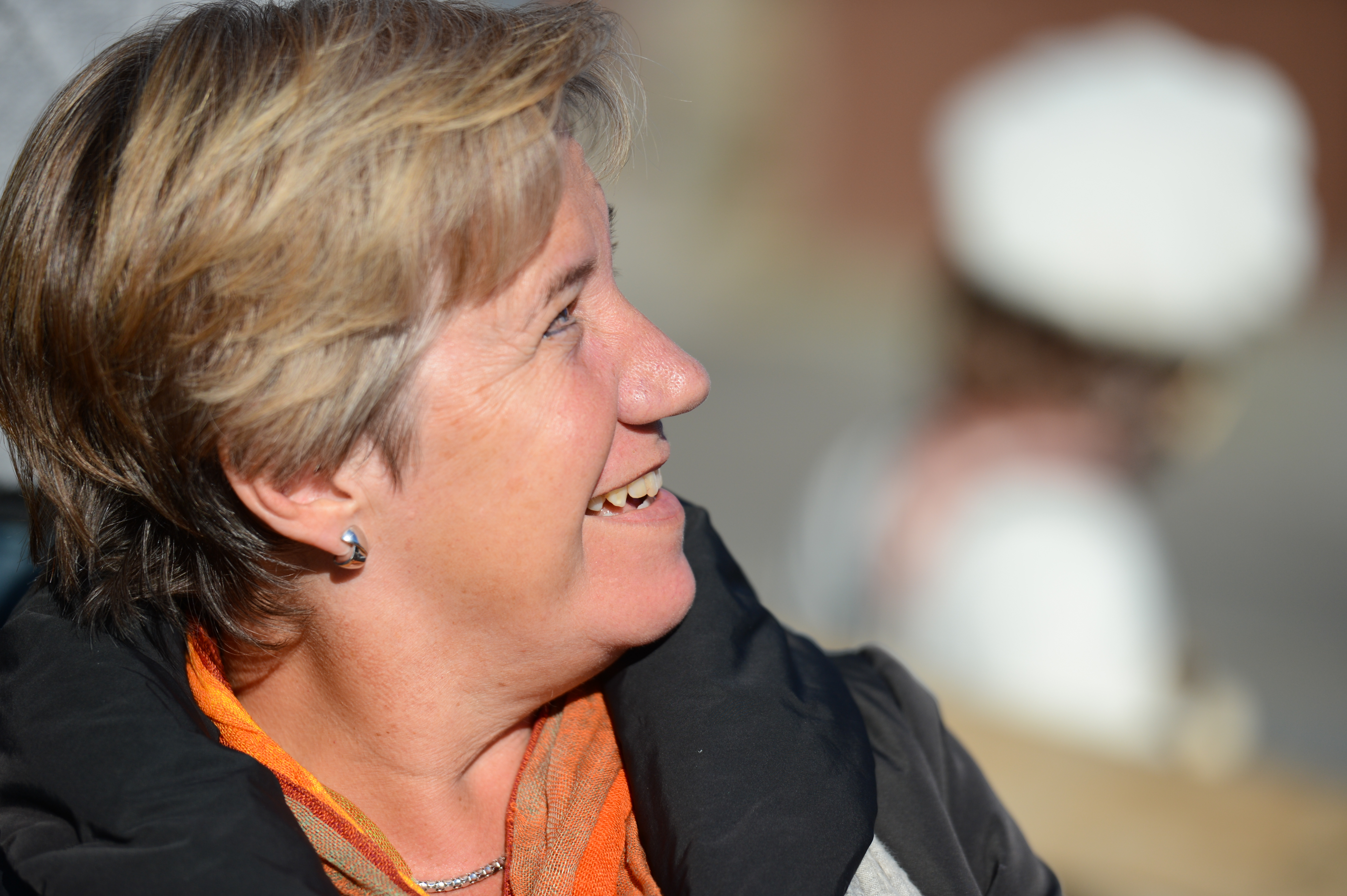 A woman with short hair and an orange scarf smiles while looking to the side.