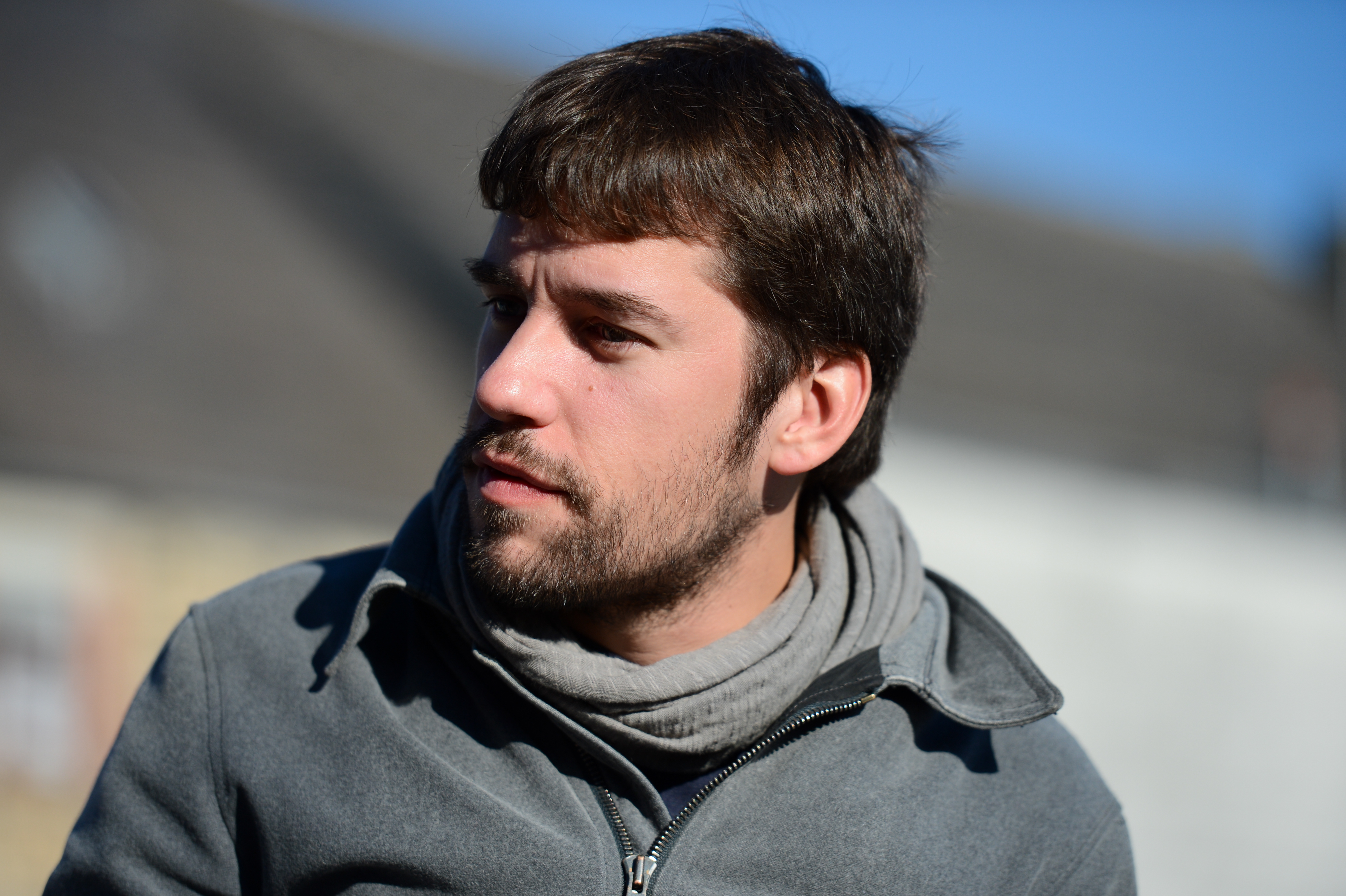 A man with short brown hair and a beard looks to the side, wearing a gray jacket and scarf outdoors.