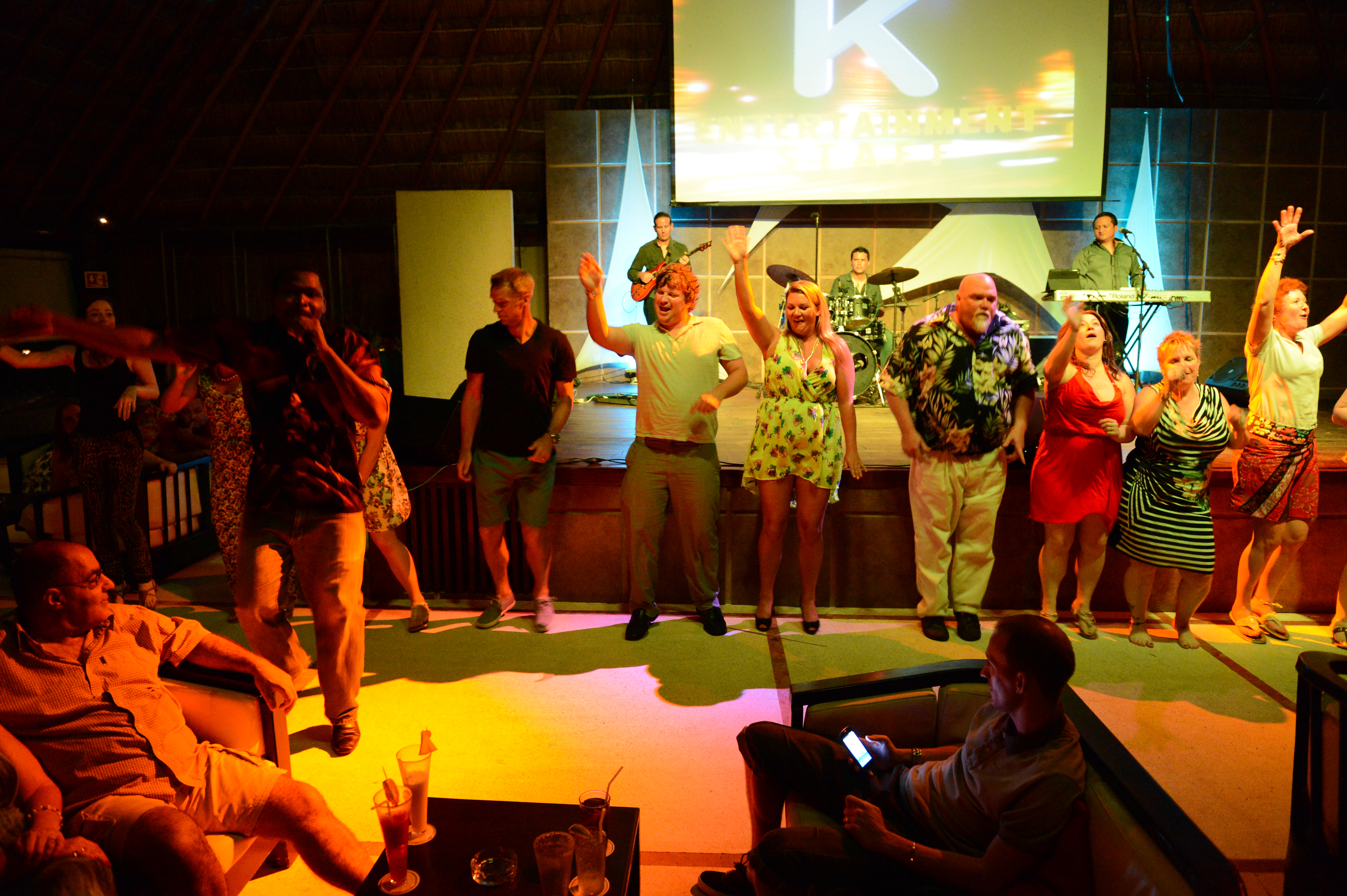 A group of people dances on stage while a live band performs in a lively indoor setting.