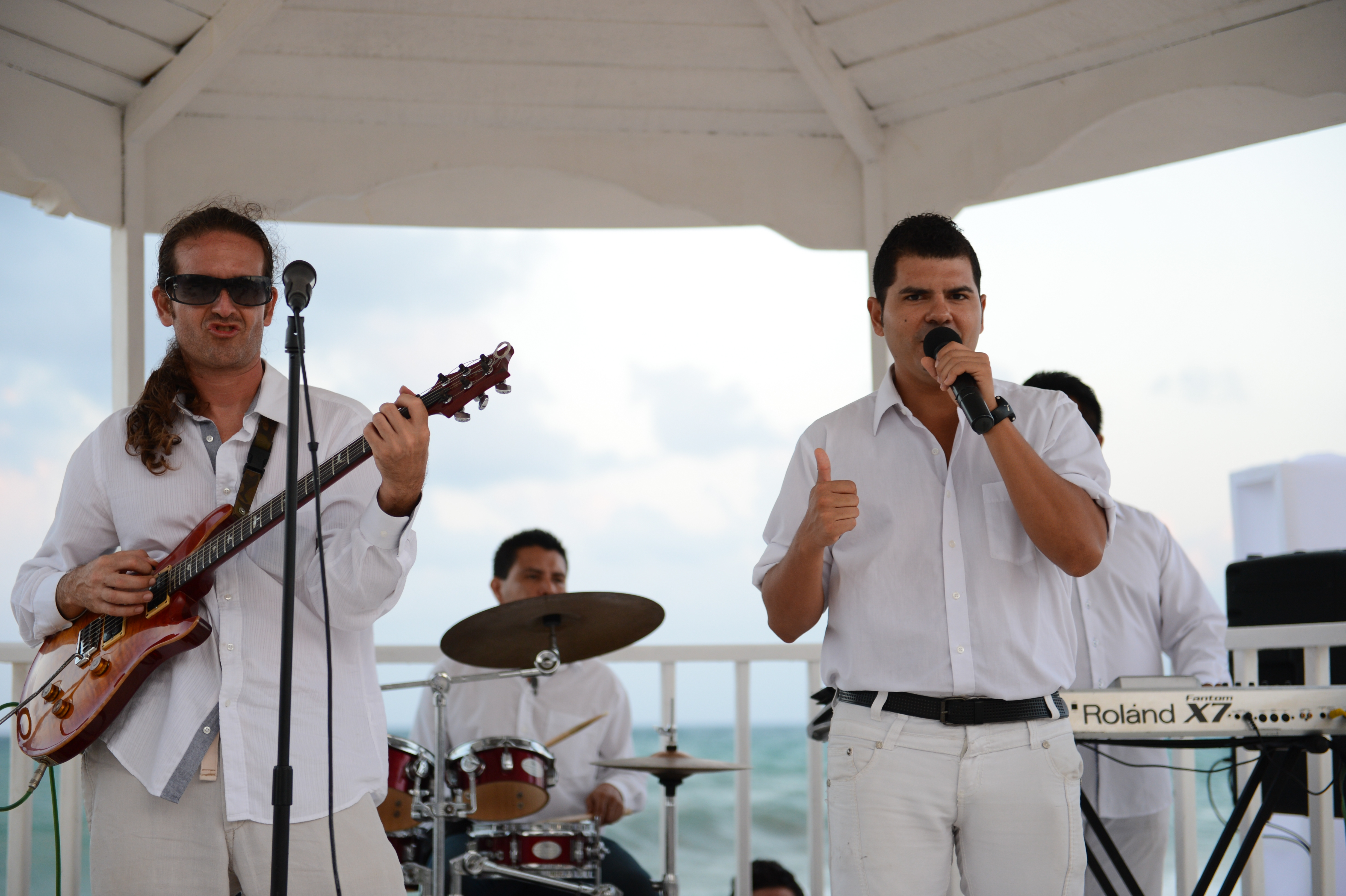 A band performs on an outdoor stage; a guitarist plays, and a singer holds a microphone while giving a thumbs-up.
