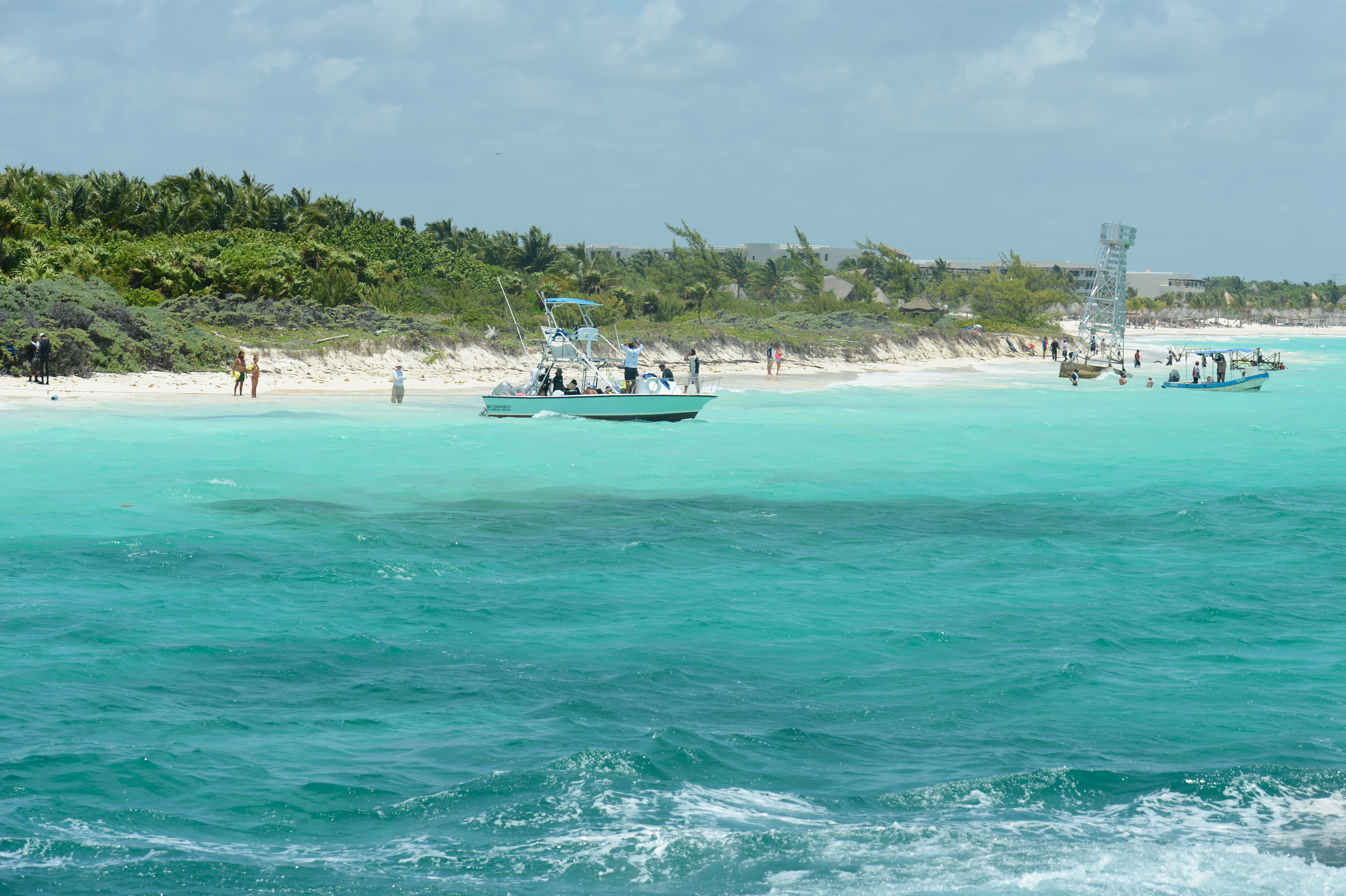 A boat is anchored near a sandy beach with people walking along the shore and in the water.
