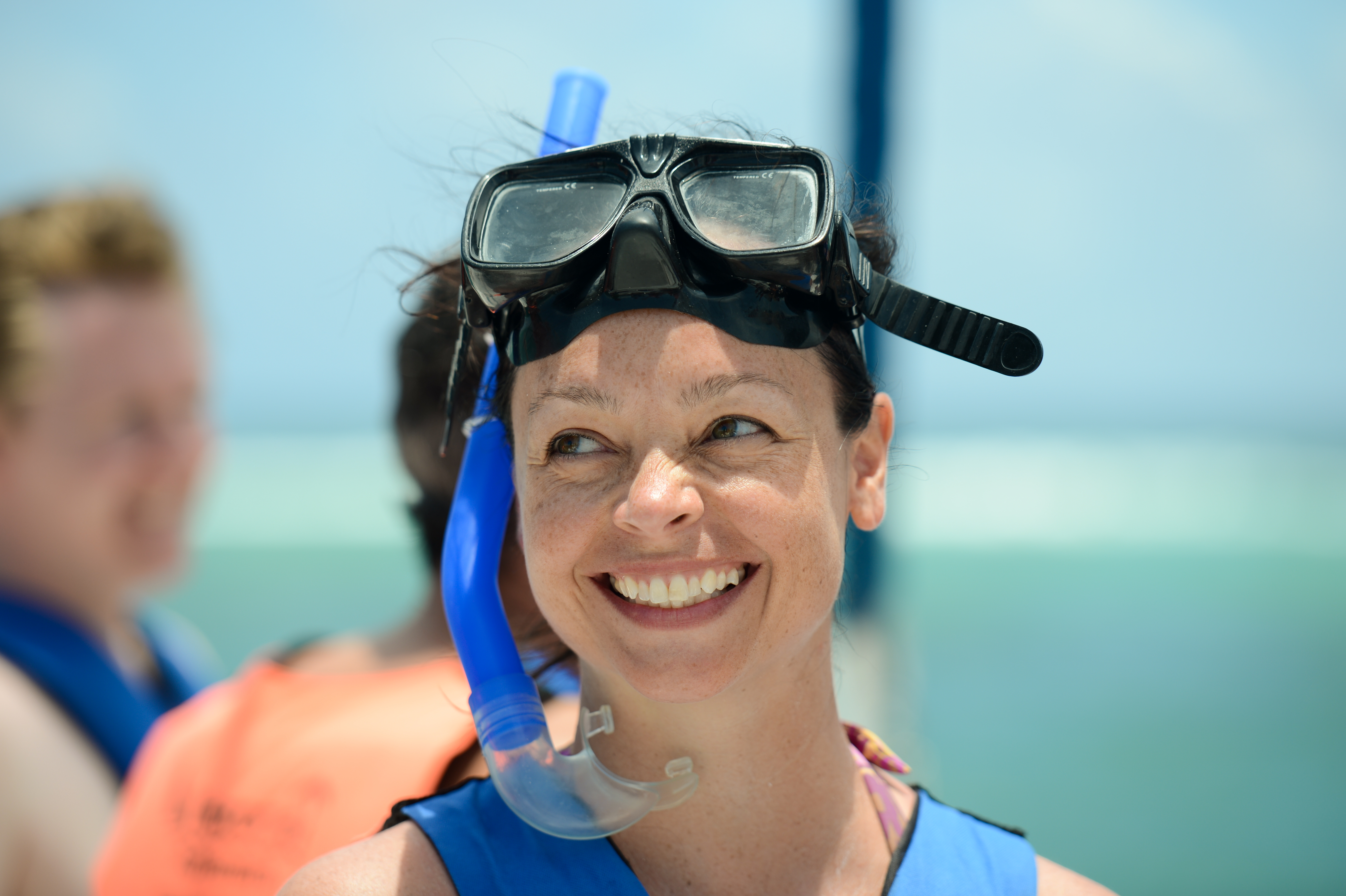 A smiling woman wearing a snorkel mask and life vest stands near the water, ready for snorkeling.