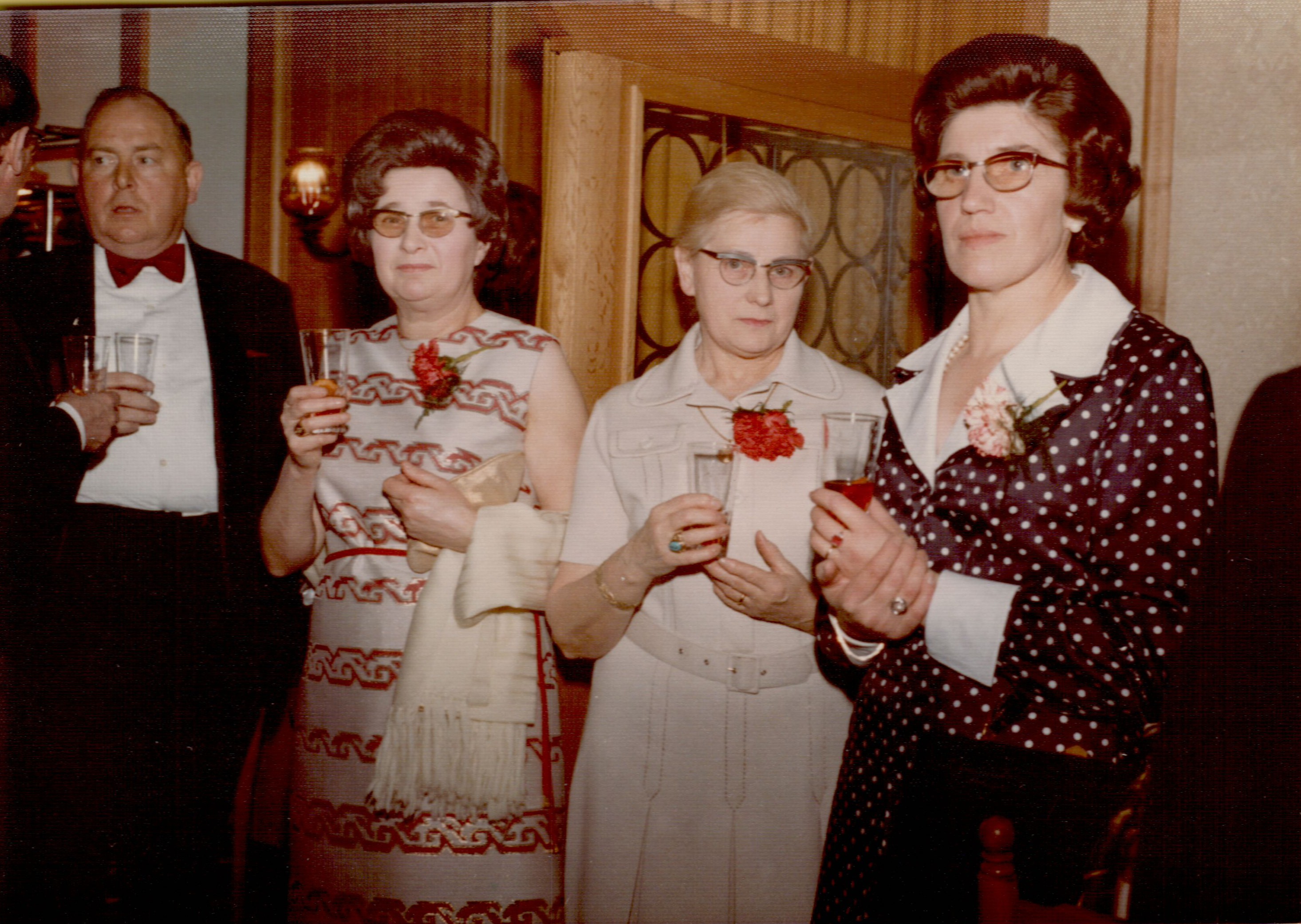 Four adults dressed formally stand together at an indoor event, holding drinks and wearing red carnation boutonnieres.