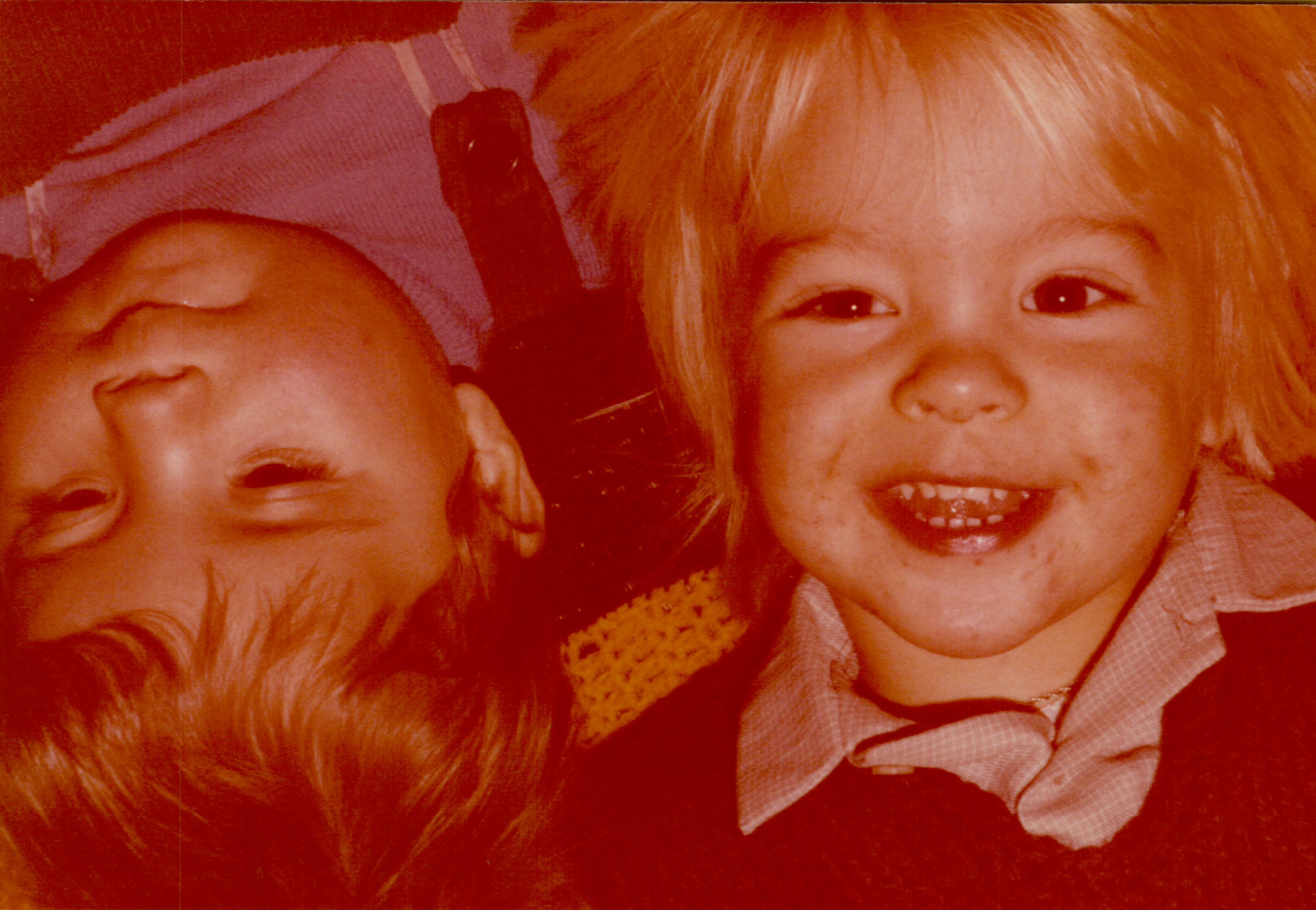 Two young children lie close together, one smiling brightly while the other rests with eyes partially closed.
