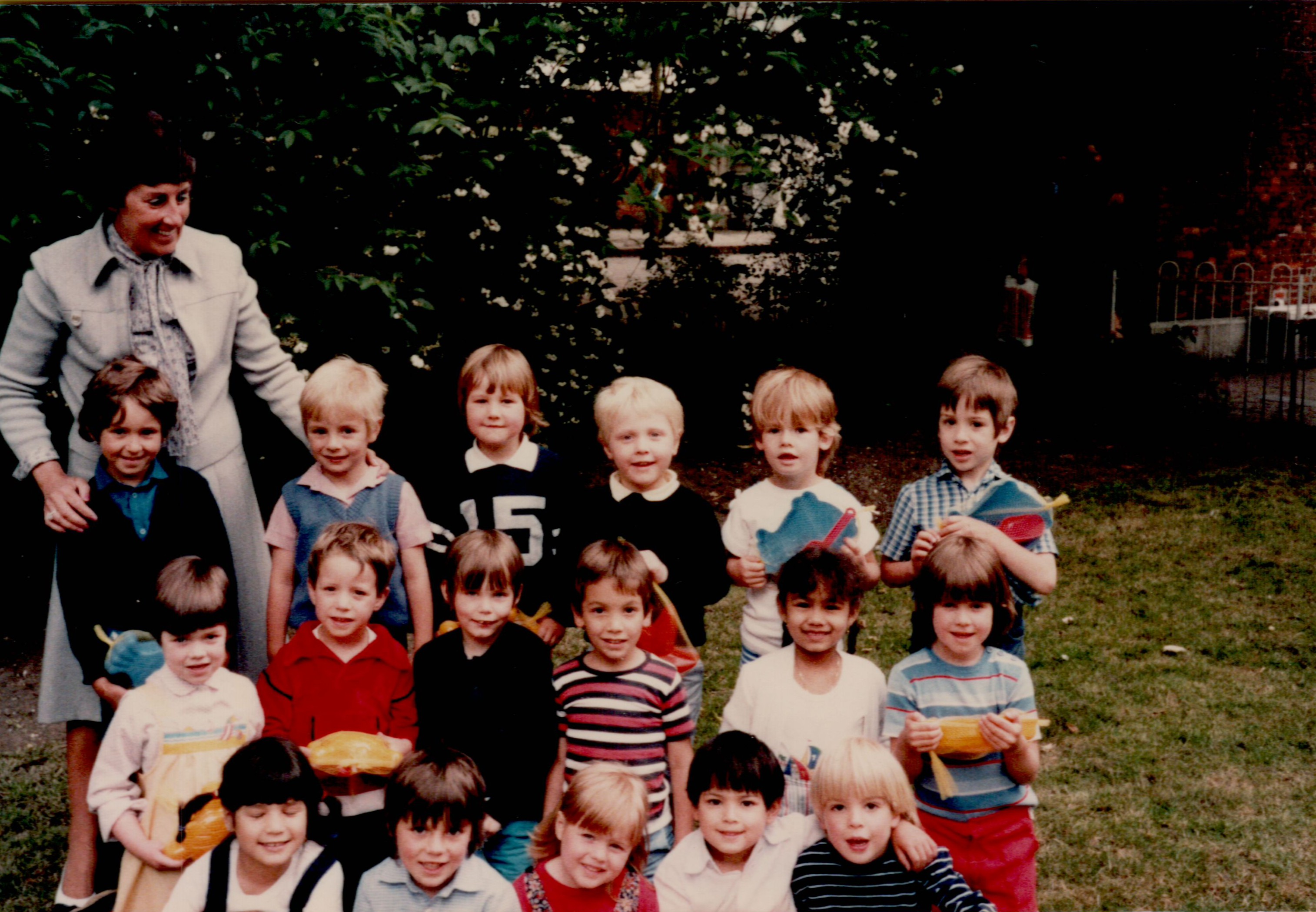 A group of young children and their teacher pose for a class photo outdoors, some holding small toys or crafts.