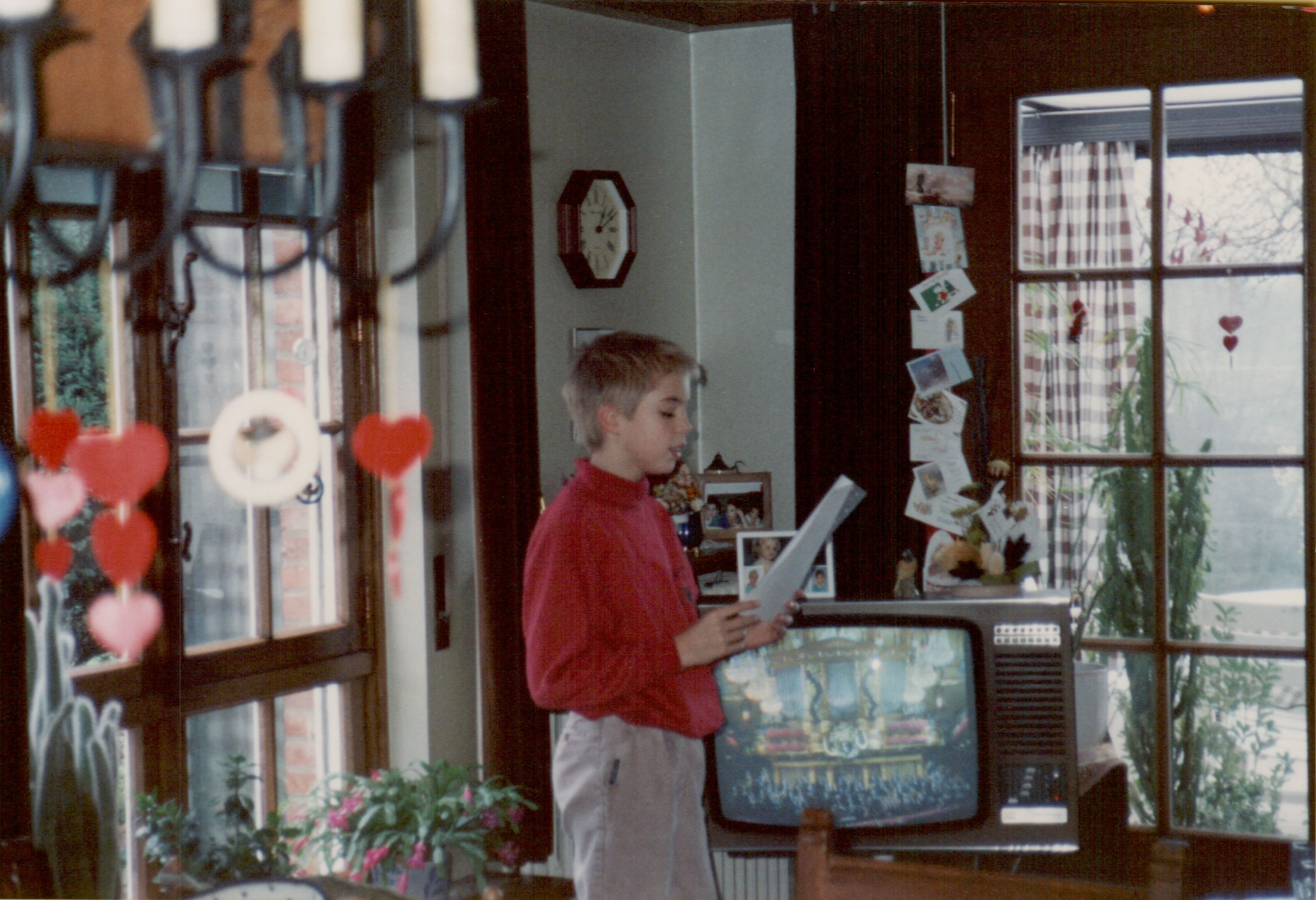 A young person in a red sweater reads a letter aloud in a decorated living room with a TV on.