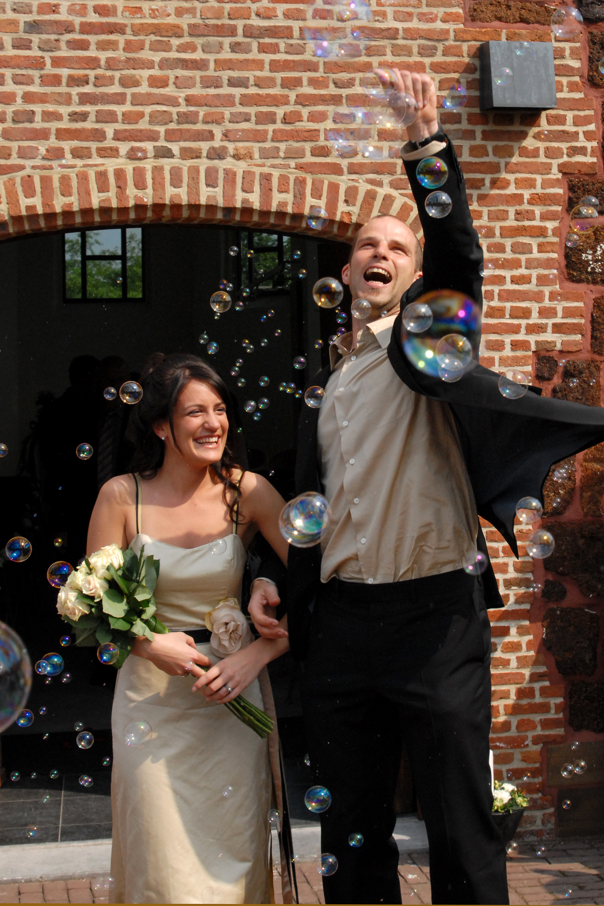 A newlywed couple smiles and celebrates outside a brick building, surrounded by floating bubbles.