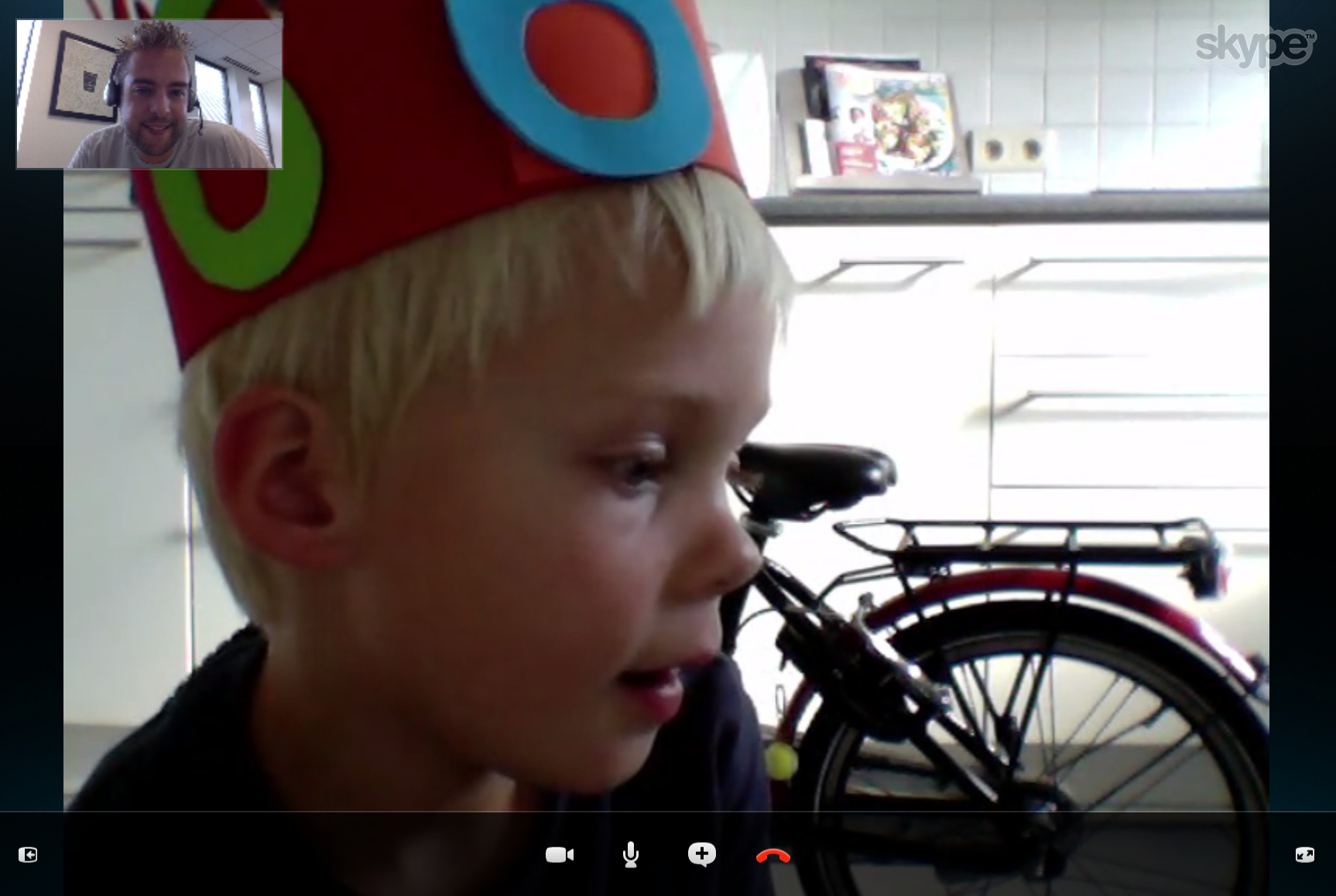 A child wearing a colorful birthday hat is on a Skype video call with a smiling adult in the corner.