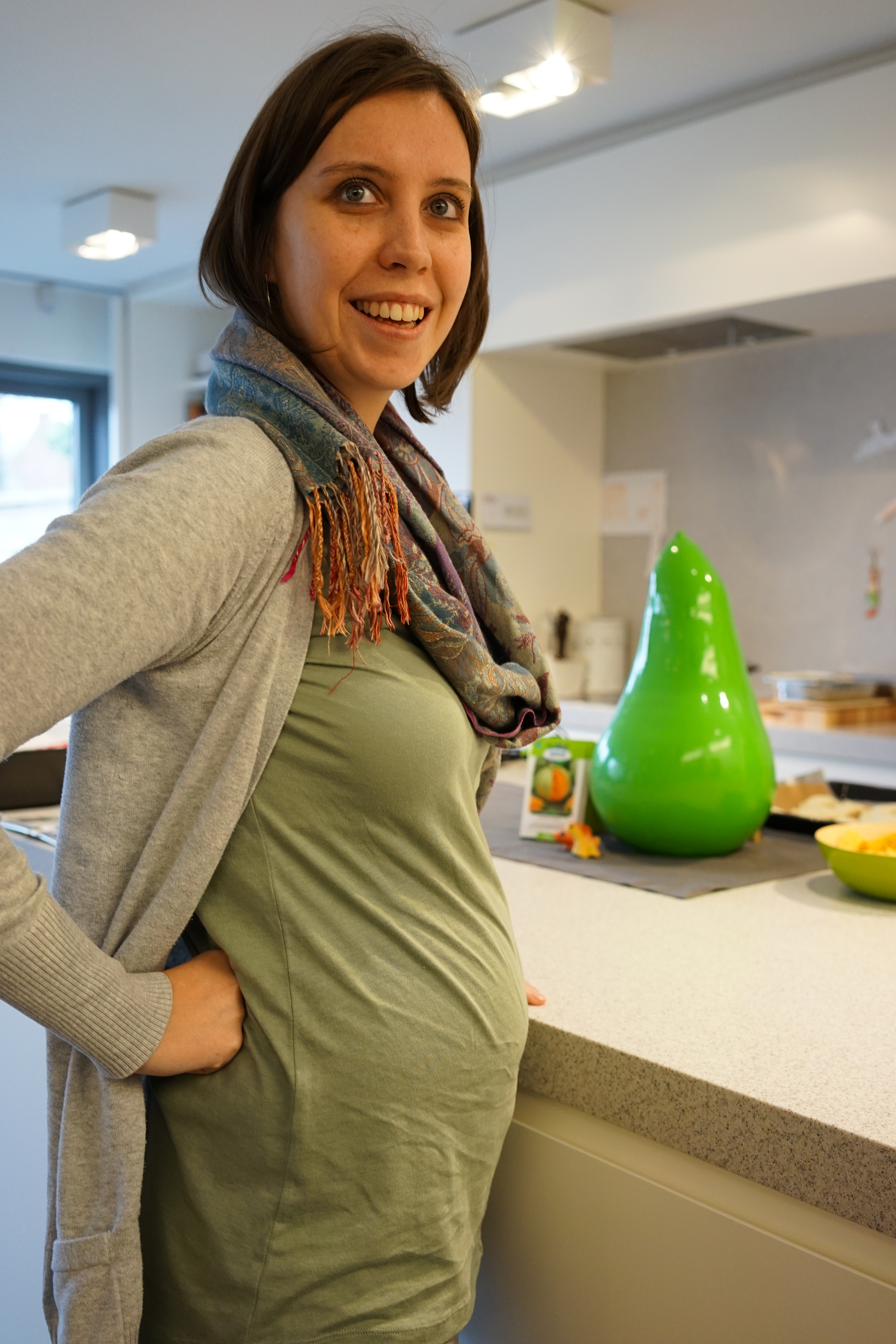 A smiling pregnant woman stands in a kitchen, resting her hand on her hip.