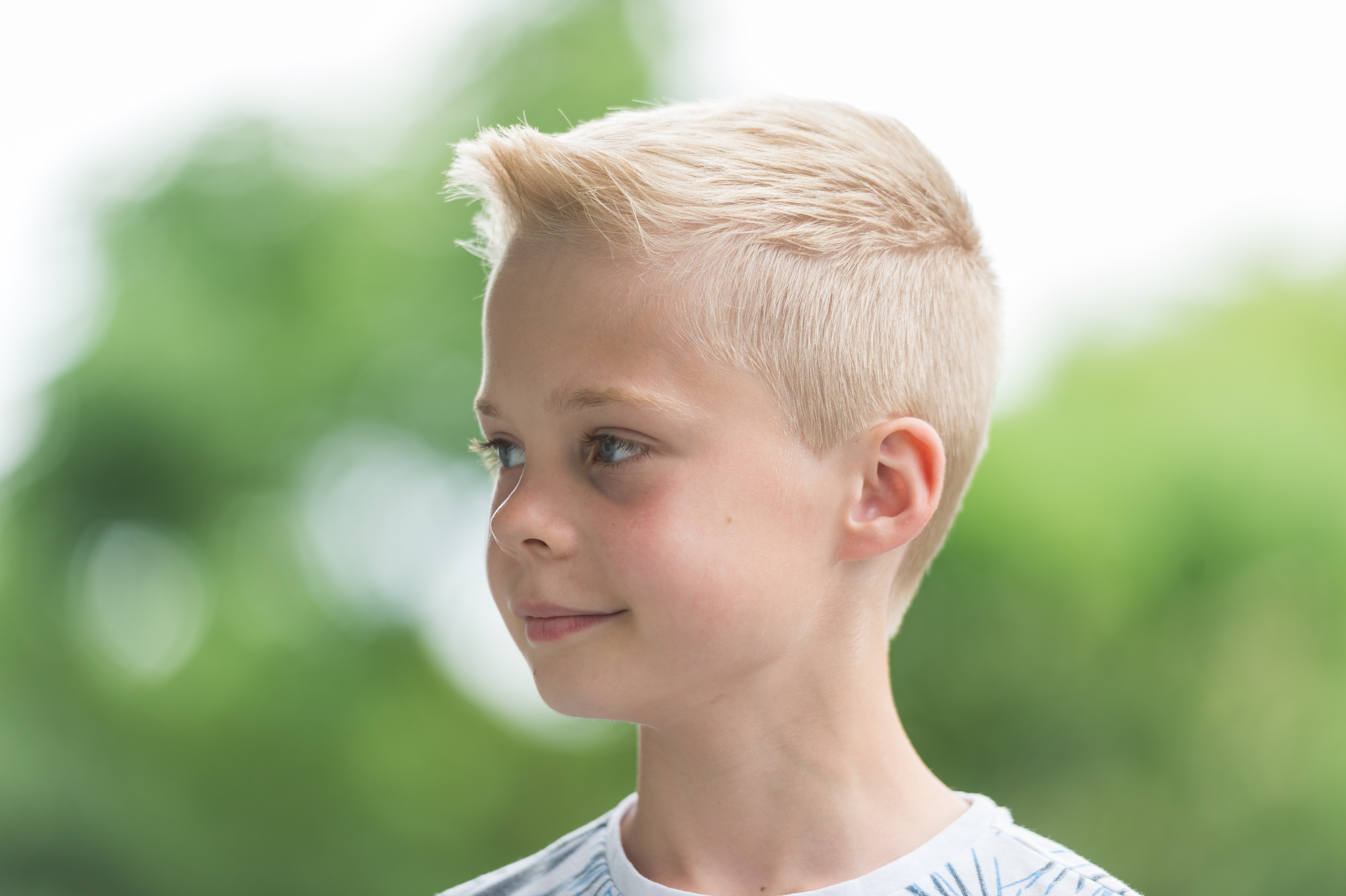 A young boy with short blond hair looks to the side with a slight smile, outdoors with a blurred green background.