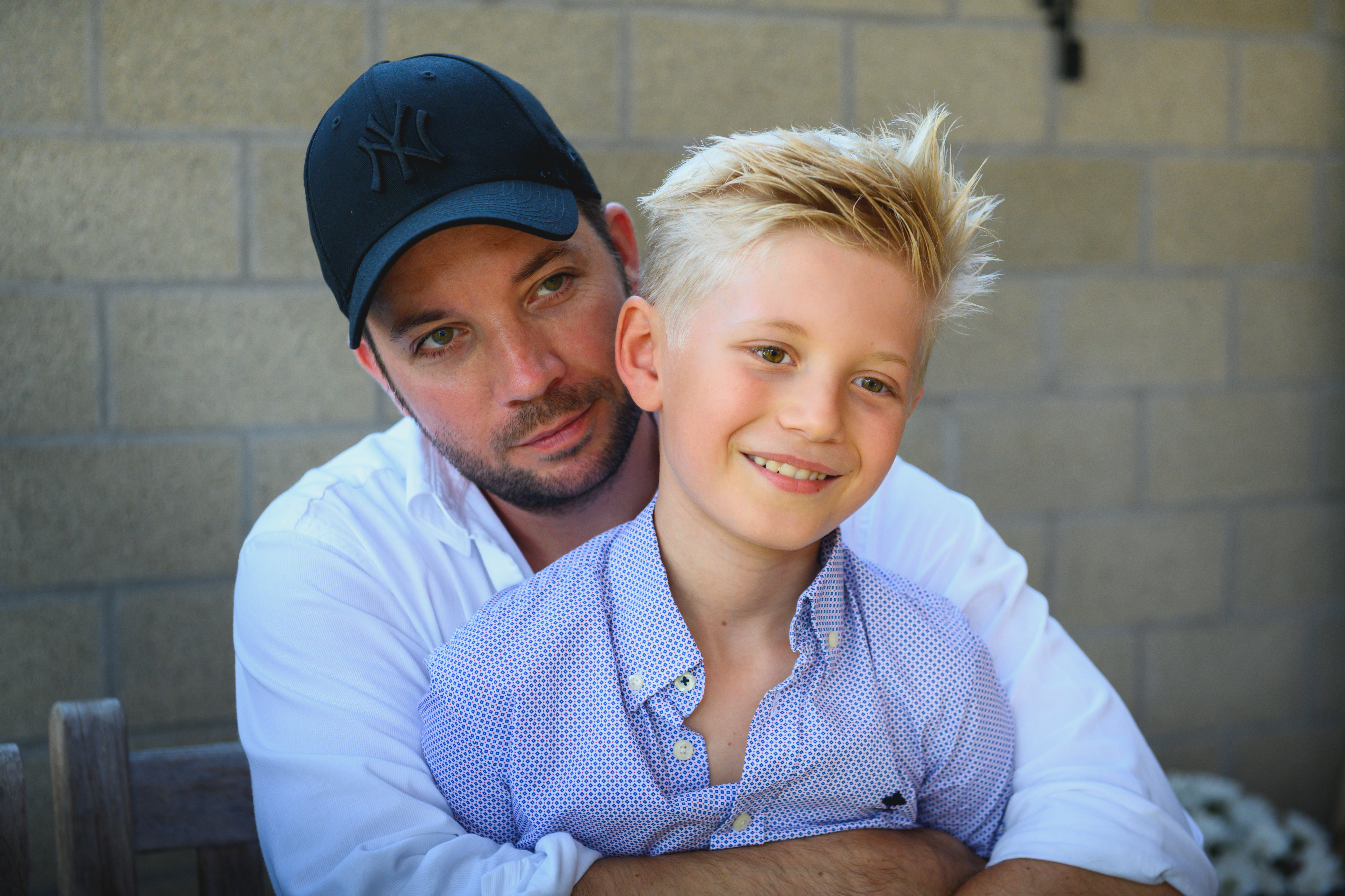 A man in a black cap hugs a smiling boy in a blue shirt while sitting outside.