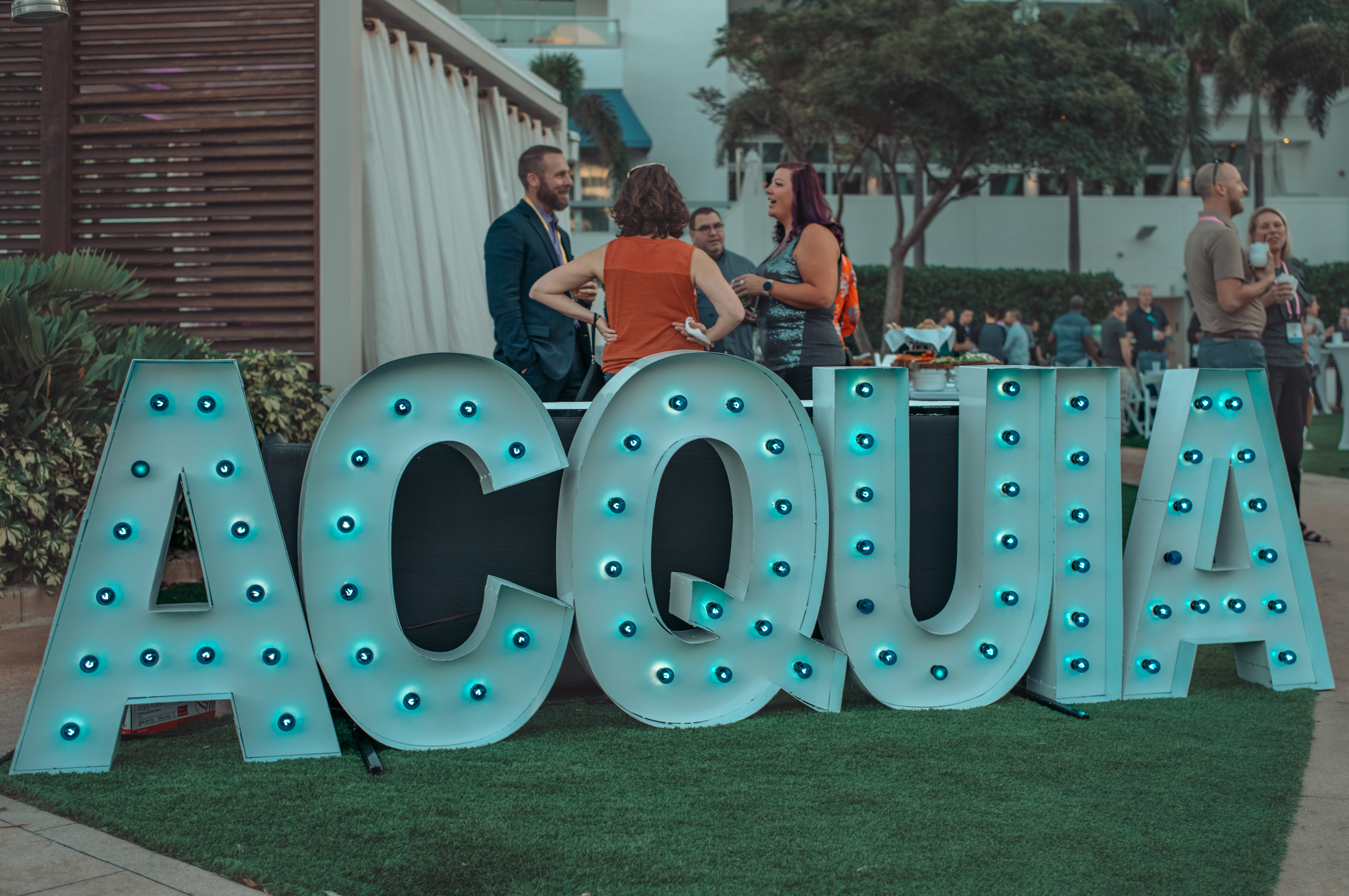 A large sign that spells out &#039;Acquia&#039; in individually lit letters. Behind the sign, people are engaged in a lively conversation.