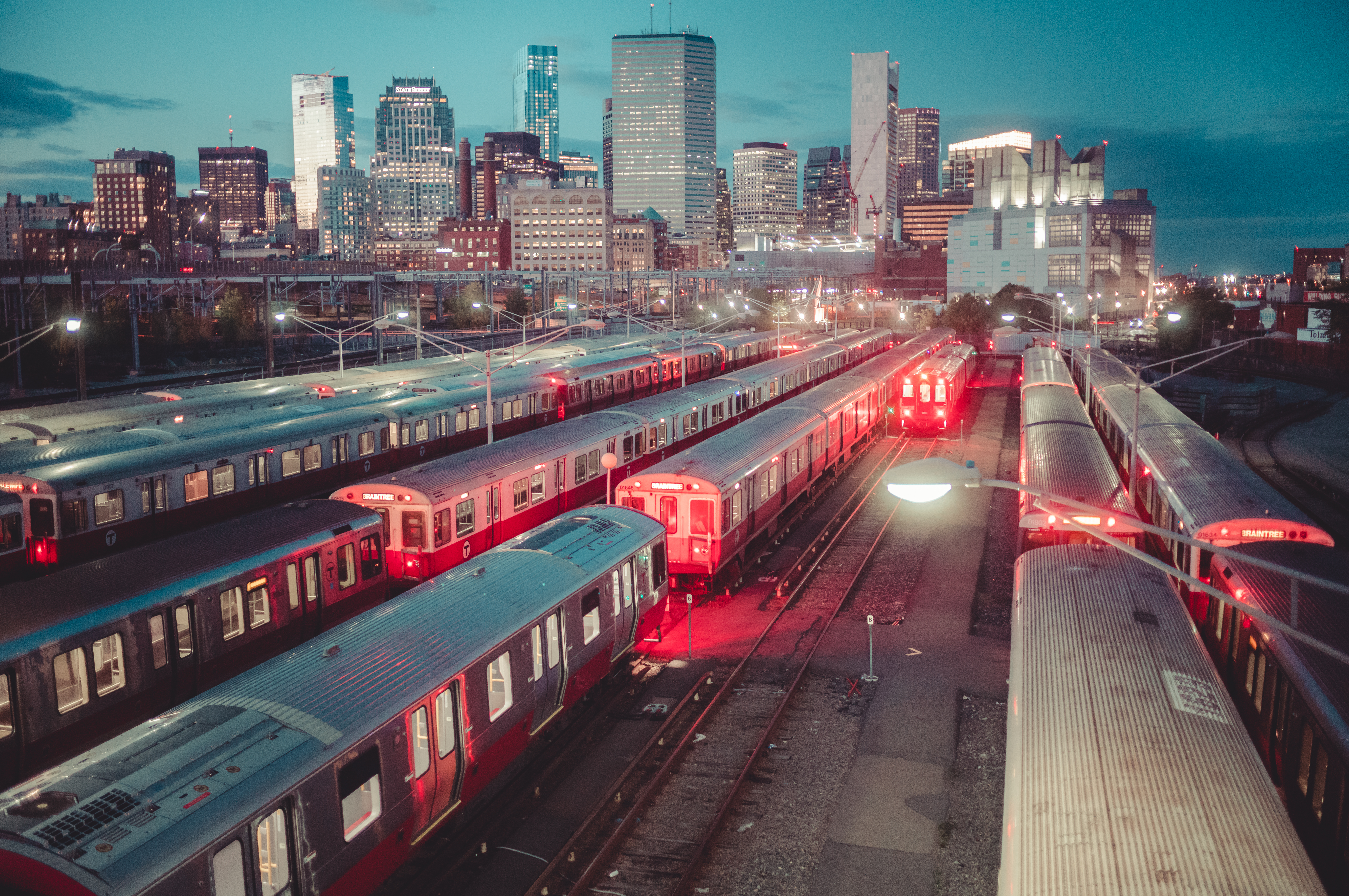 Multiple red subway trains are parked in a train yard near Boston&#039;s South Station, with the city skyline behind them.