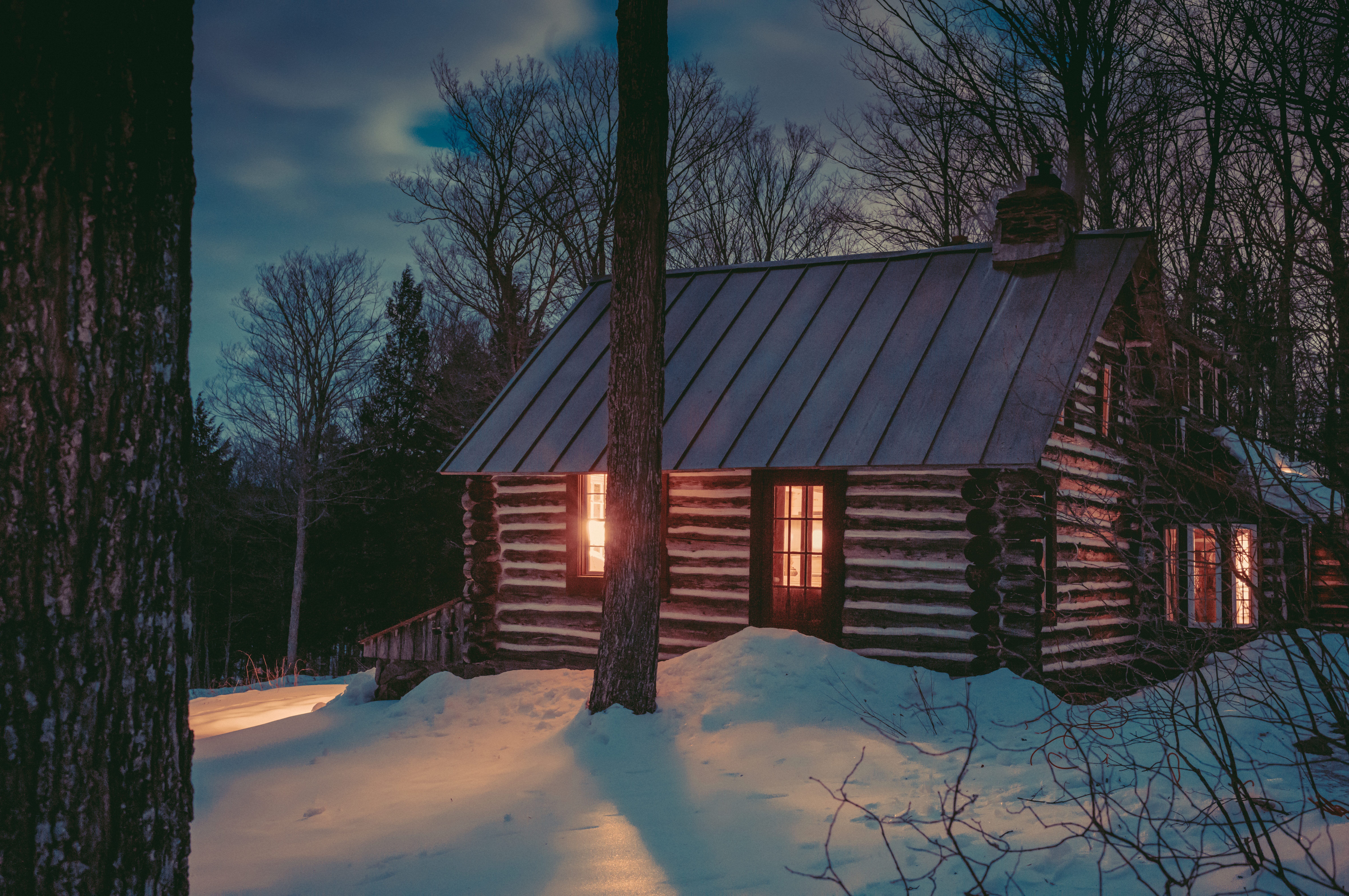 A log cabin nestled among trees, set in the evening&#039;s darkness, its interior lights reflecting on the snow and creating a peaceful atmosphere.