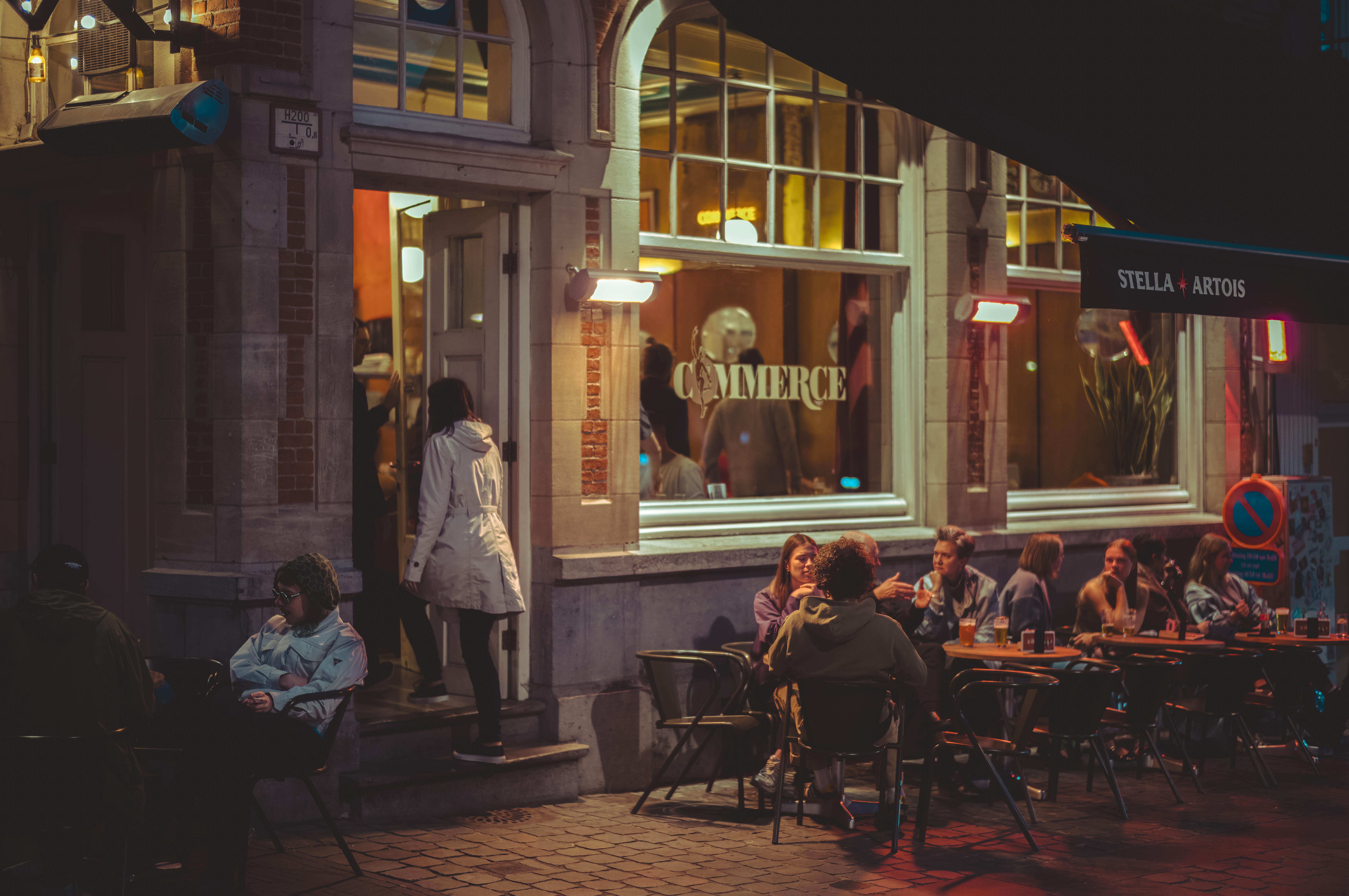 A nighttime view of a bar with &quot;Commerce&quot; printed on its windows, people drinking at outdoor tables, and a person entering the doorway.