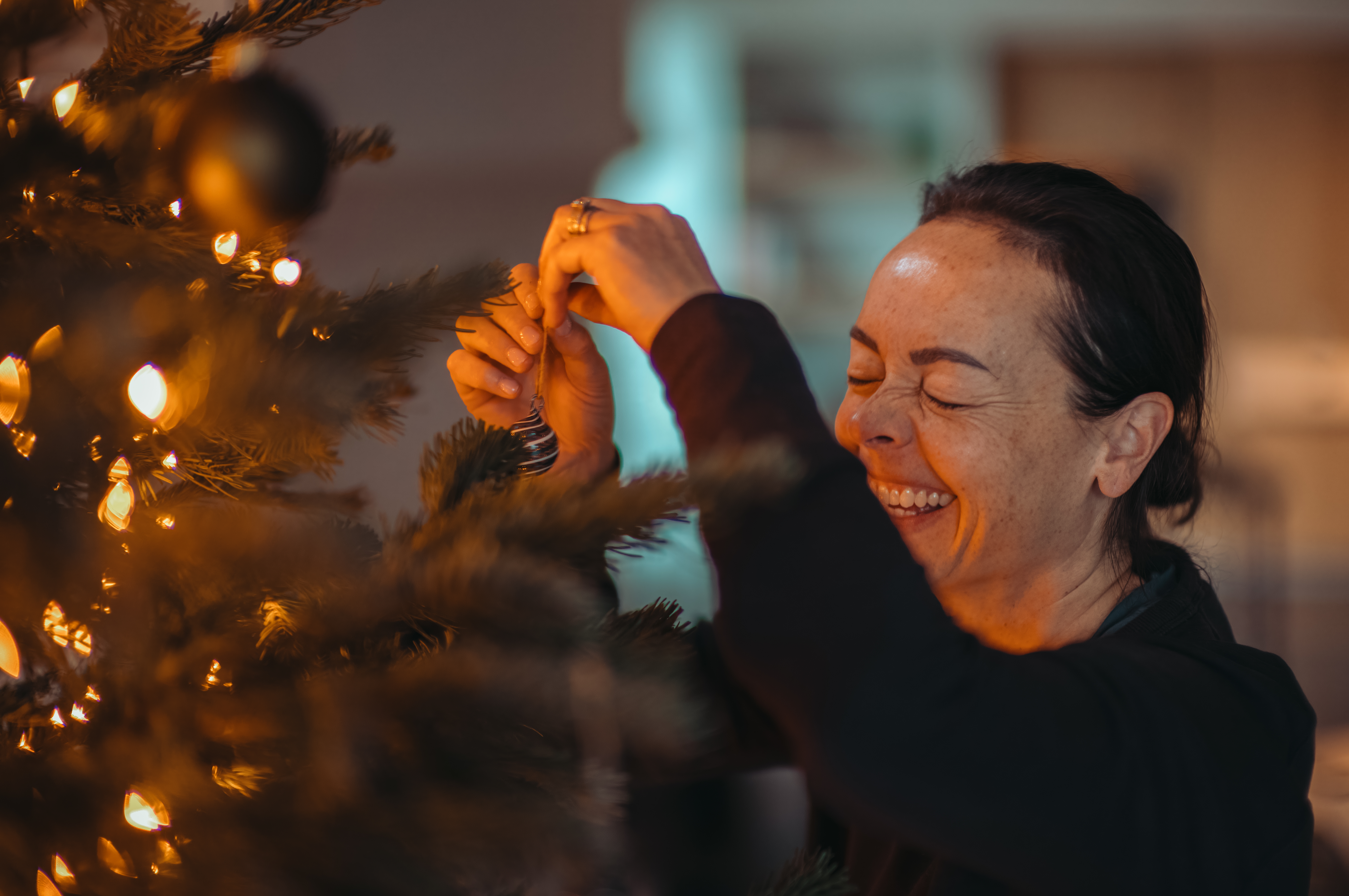 A woman laughing as she hangs an ornament on a Christmas tree.