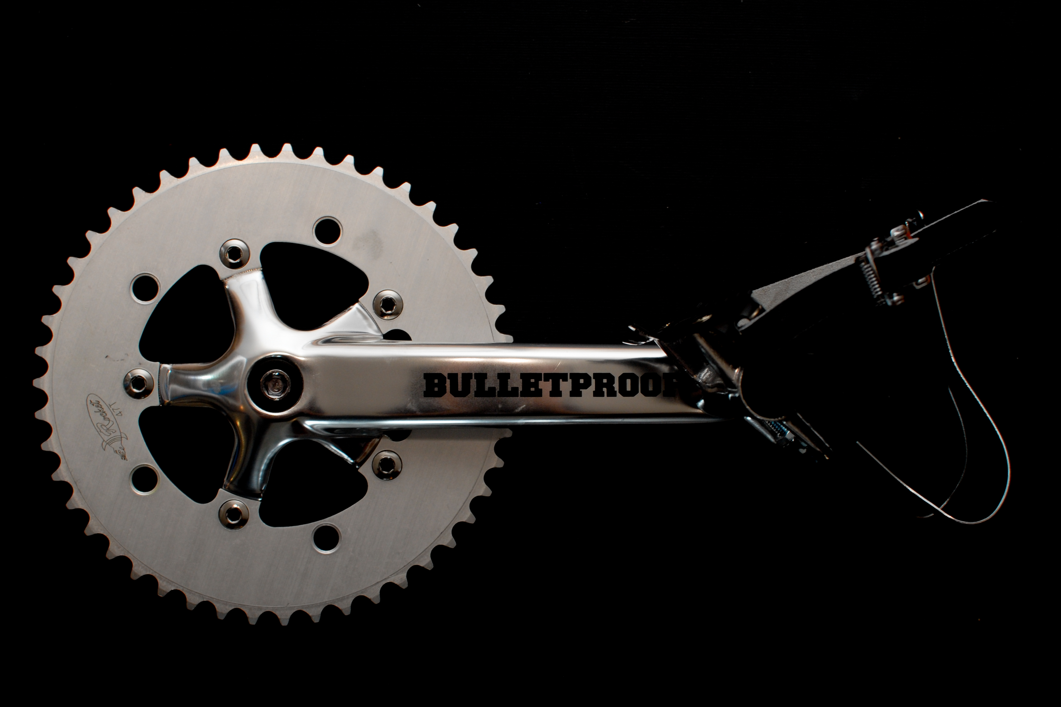 Close-up of a bicycle crankset with &quot;Bulletproof&quot; printed on the crank arm, set against a dark background.