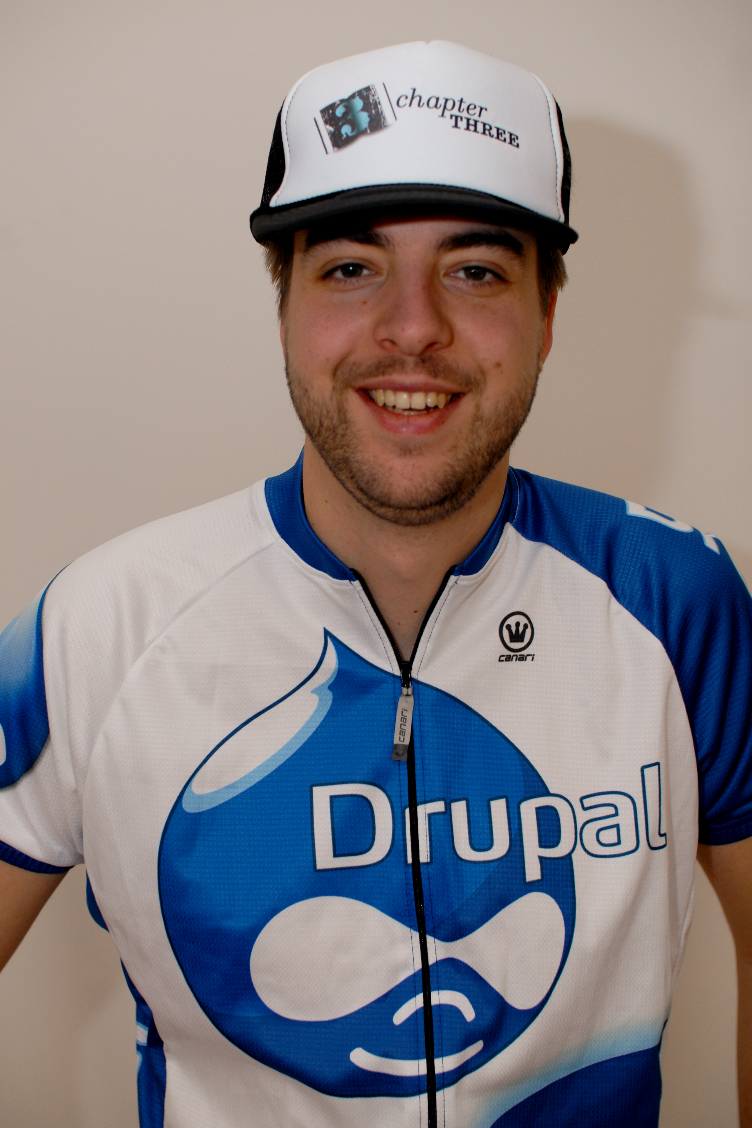 A smiling person wears a Drupal cycling jersey and a cap with &quot;Chapter Three&quot; printed on it.