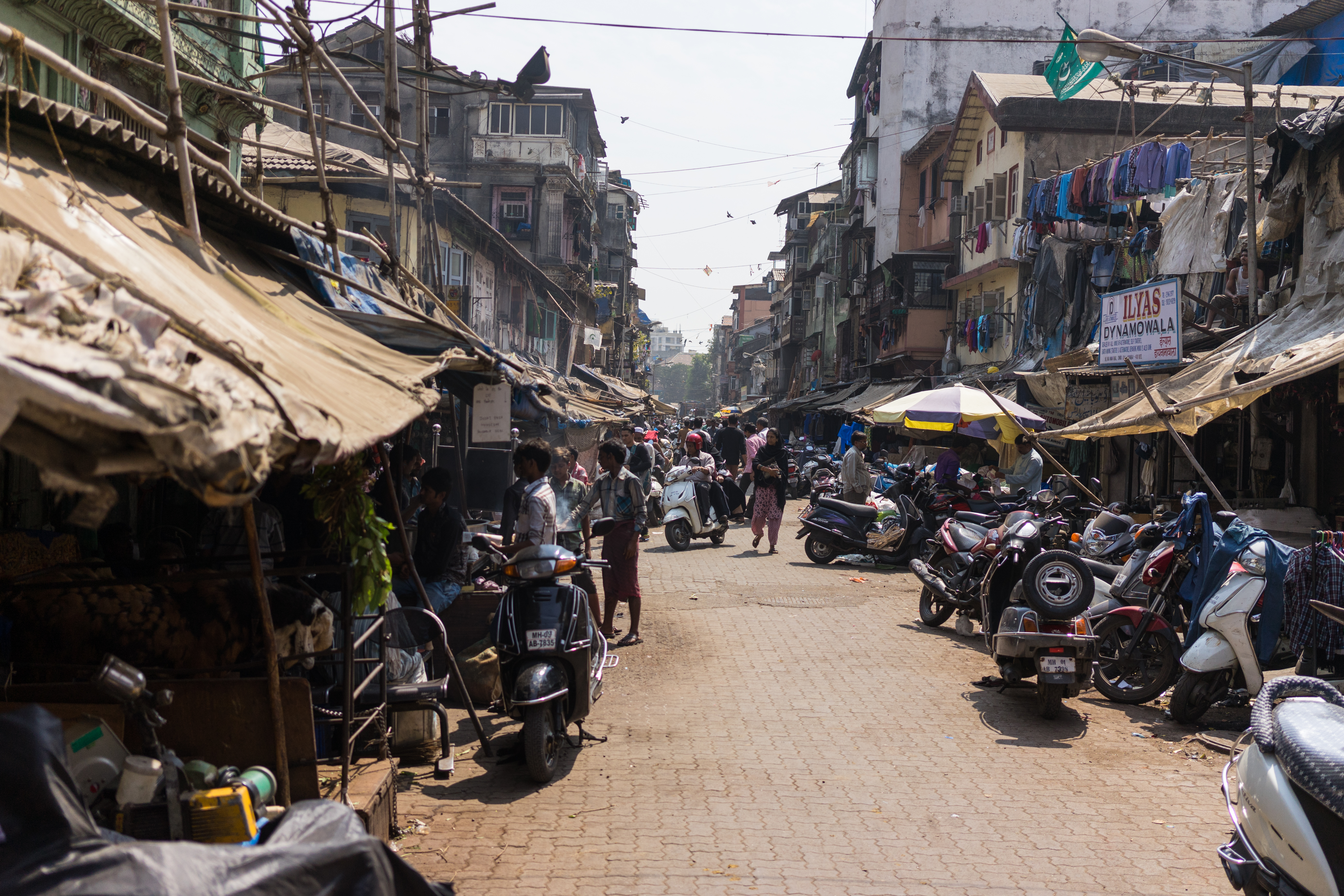 A busy street market with people walking, scooters parked, and vendors selling goods under makeshift stalls.