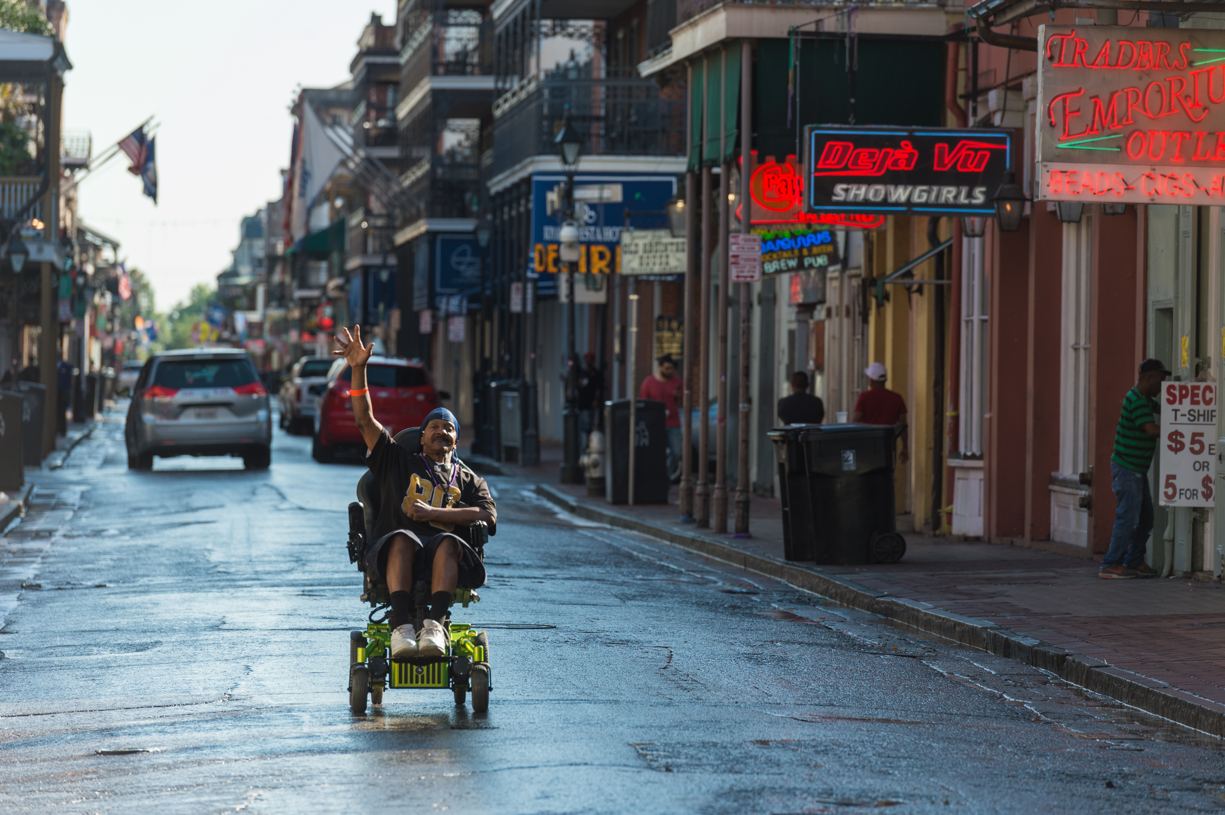 A person in a motorized wheelchair moves down a wet street, raising one hand while wearing a sports jersey.