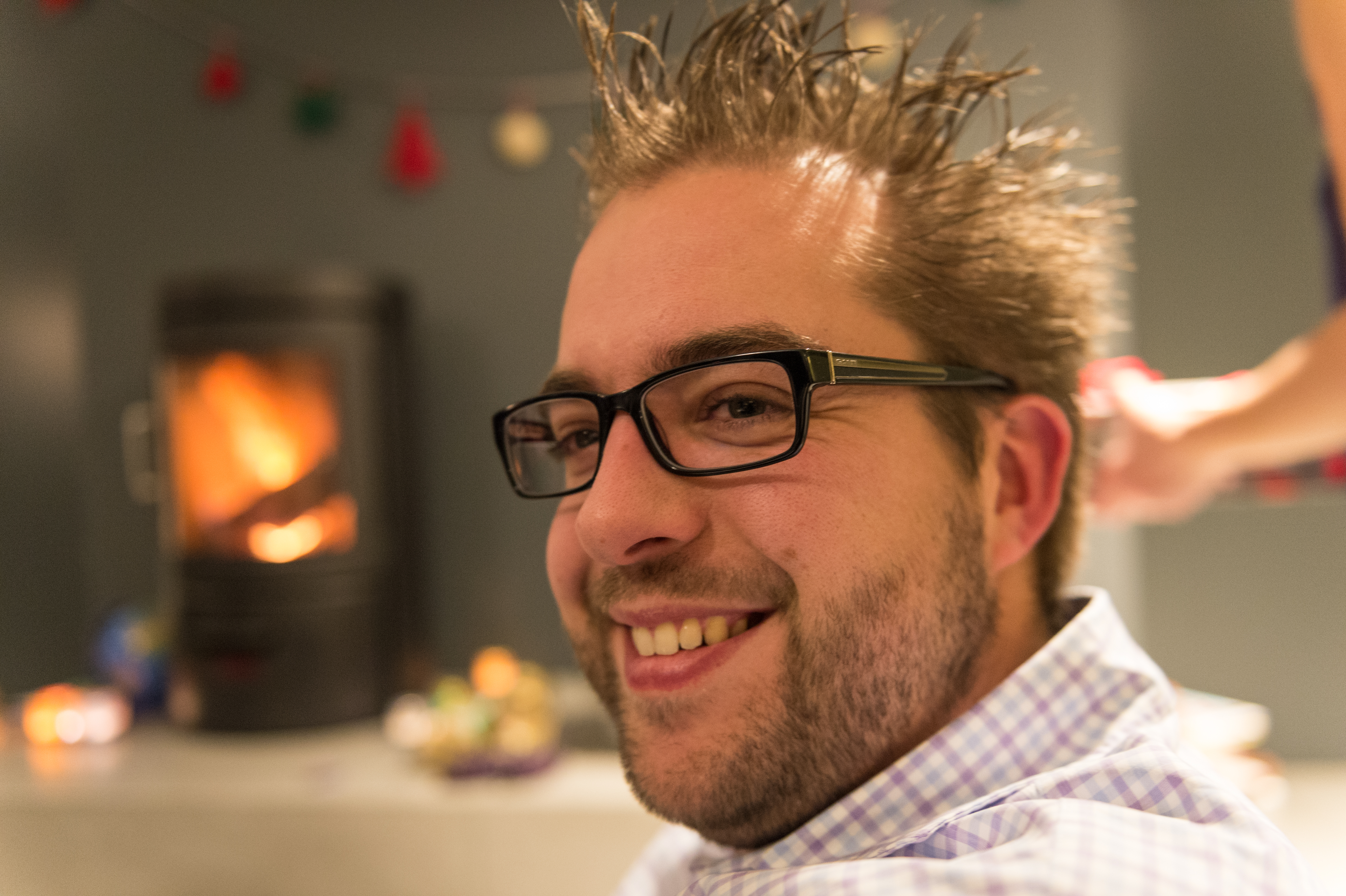 A smiling man with glasses and spiky hair sits indoors while someone styles his hair in the background.