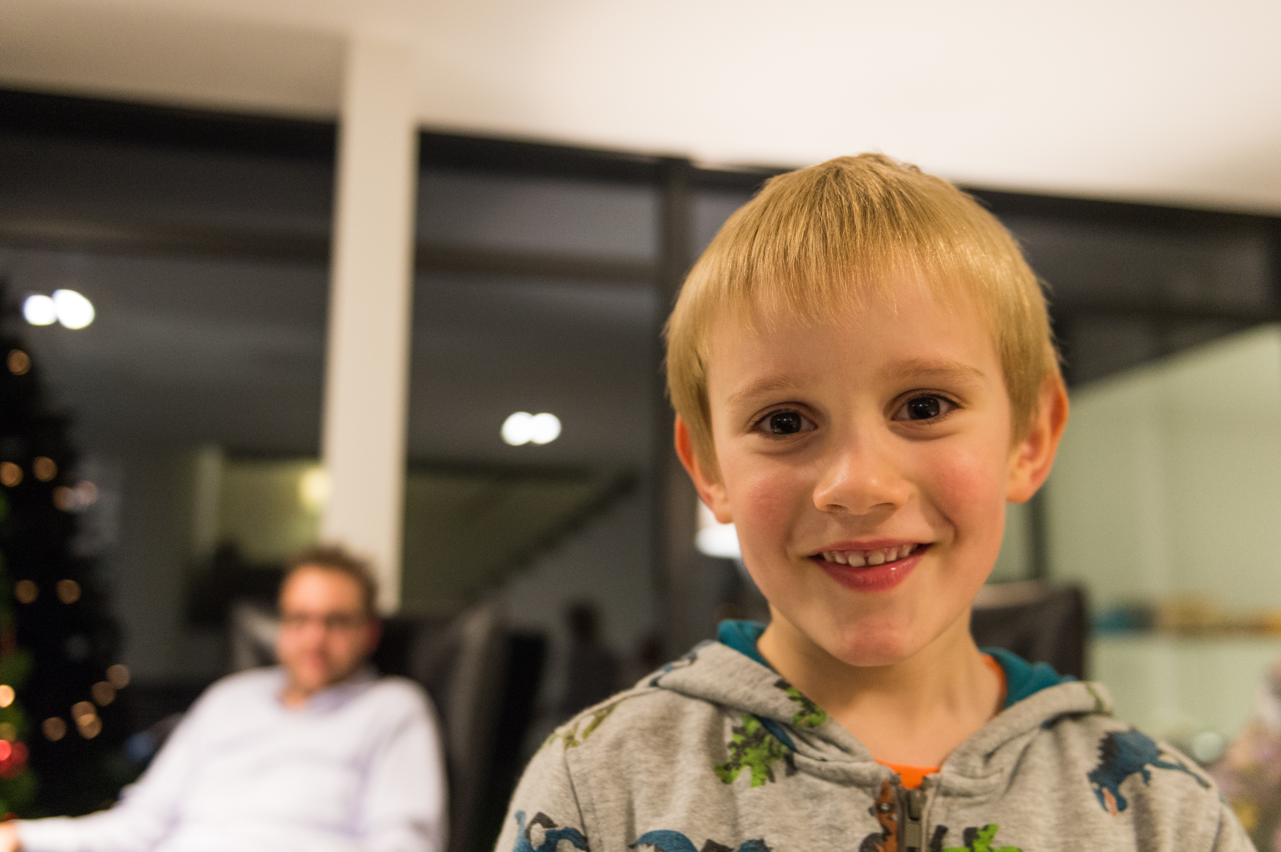 A smiling child in a dinosaur hoodie stands in the foreground, with an adult sitting in the background.