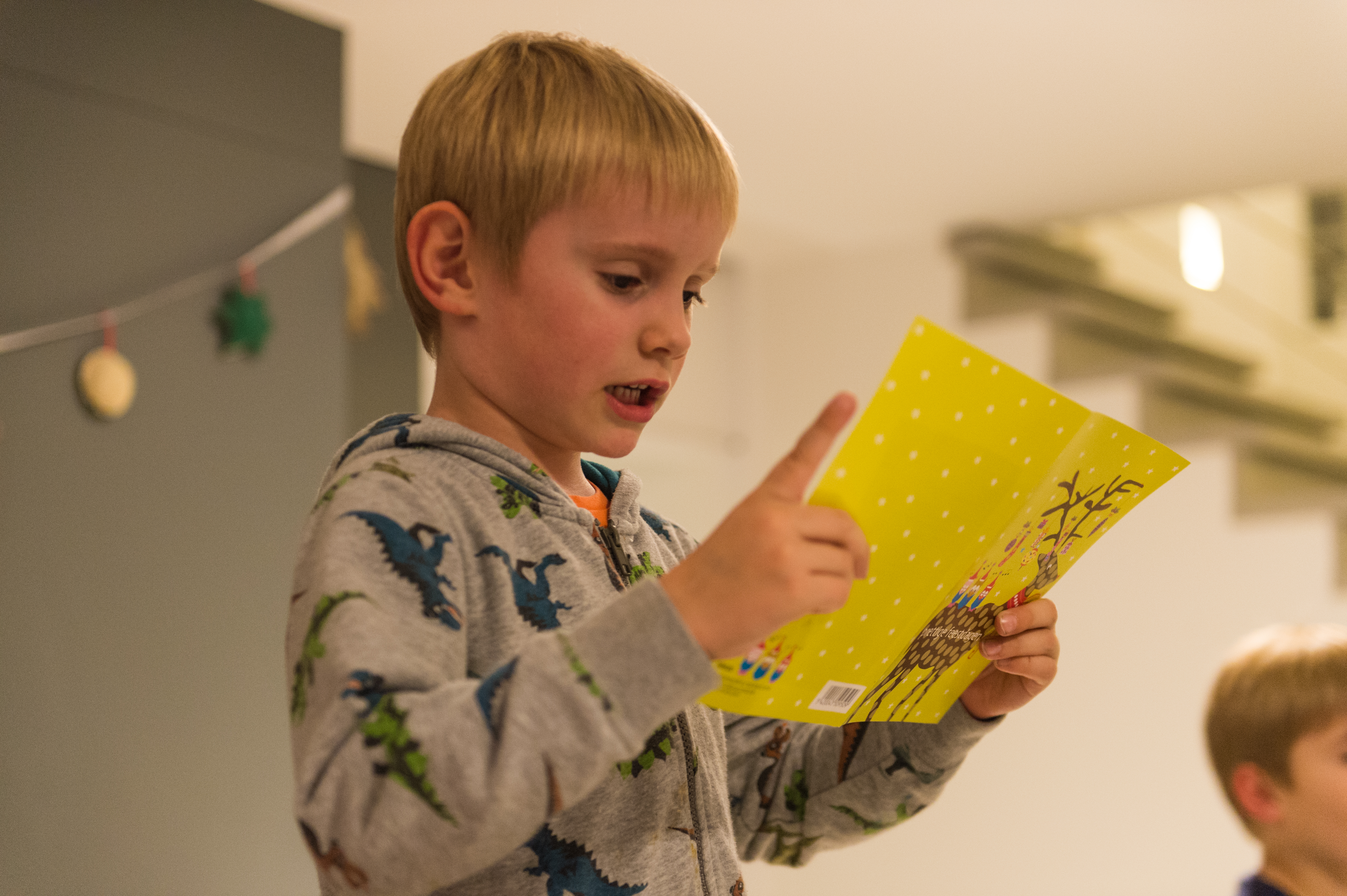 A child in a dinosaur hoodie reads from a yellow card, gesturing with one finger during a New Year's Eve party.