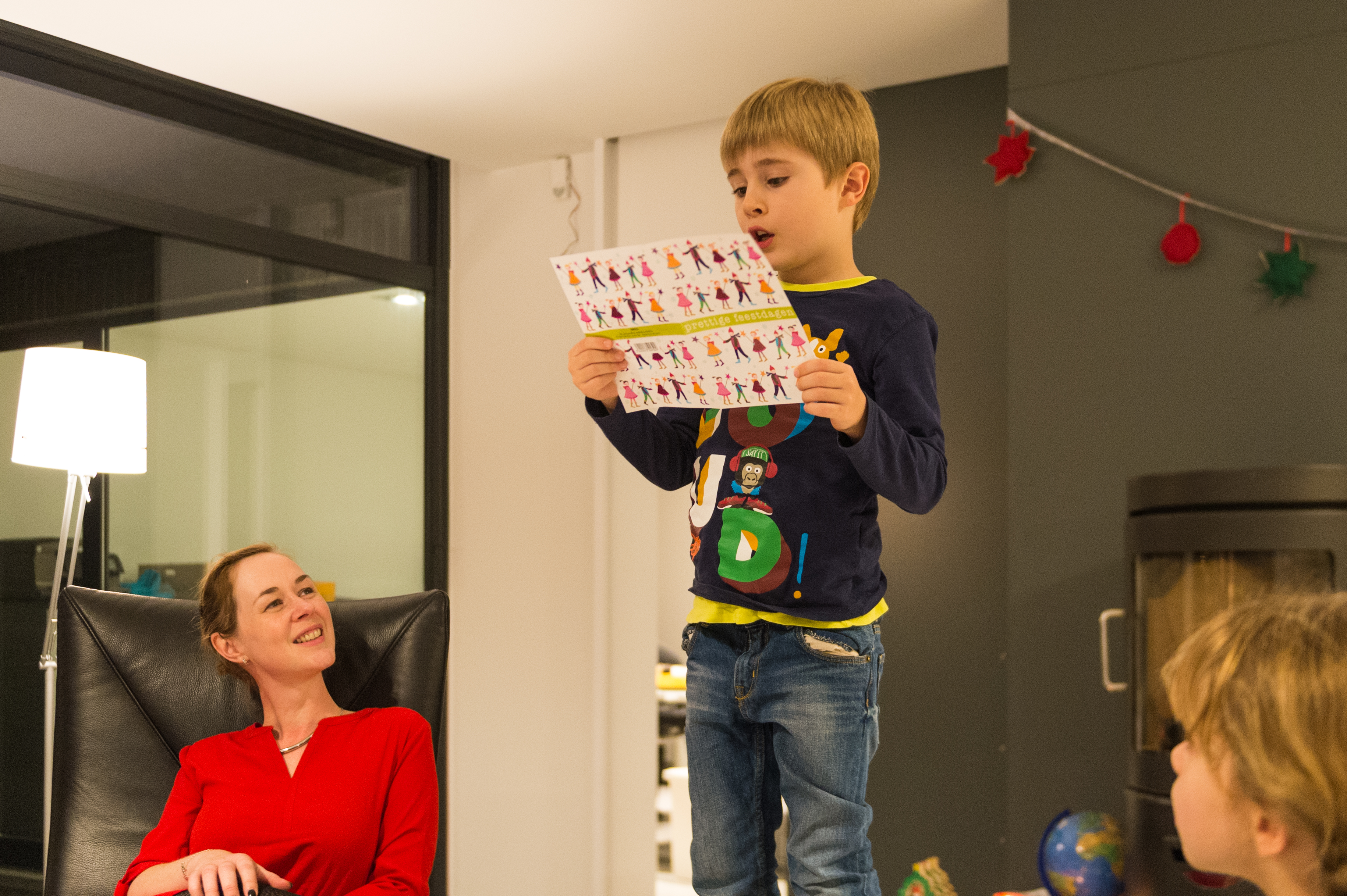 A young boy stands on a chair, reading from a colorful paper, while others watch and smile.