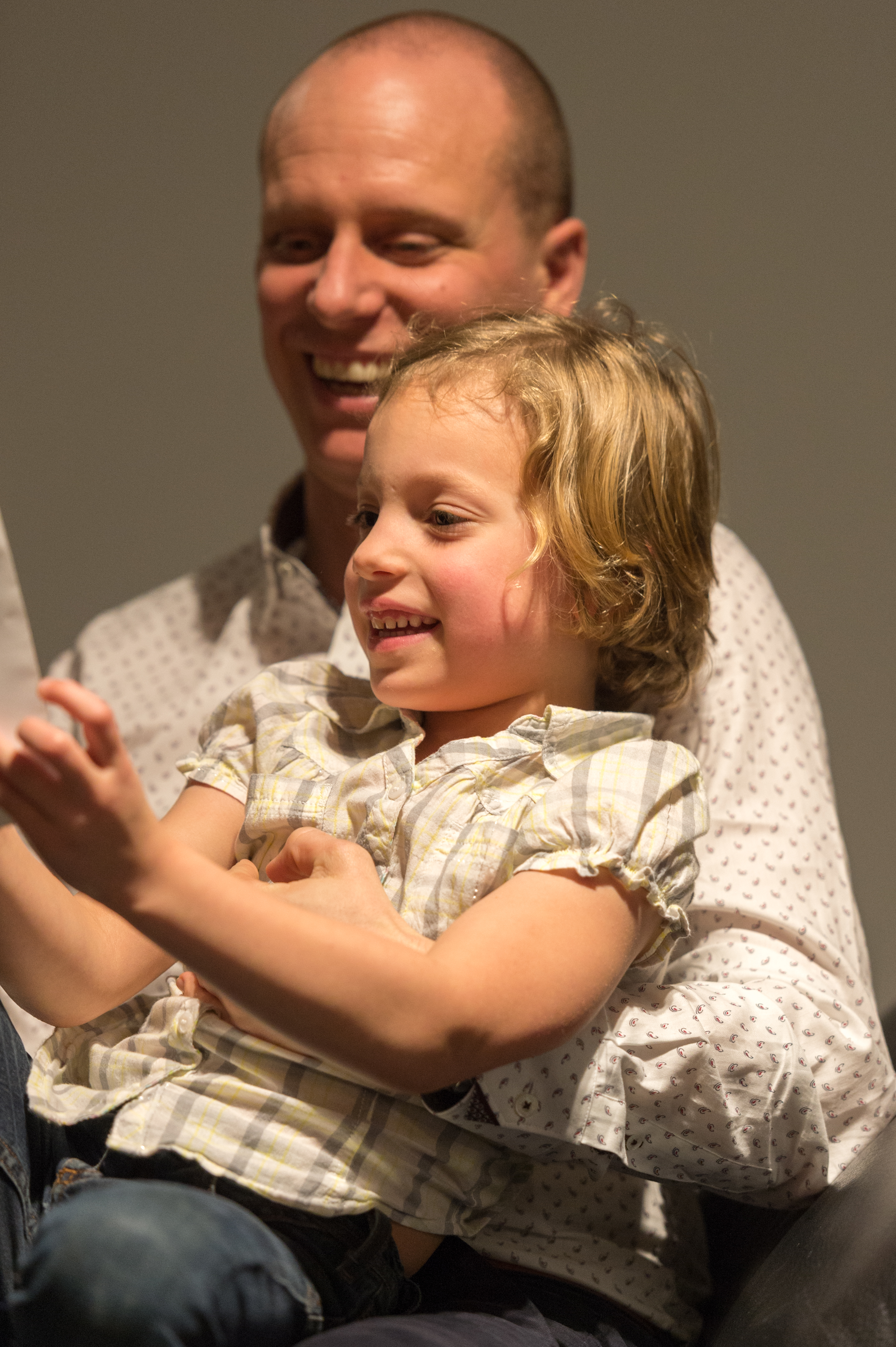 A smiling child sits on an adult's lap, holding something and looking at it with excitement.