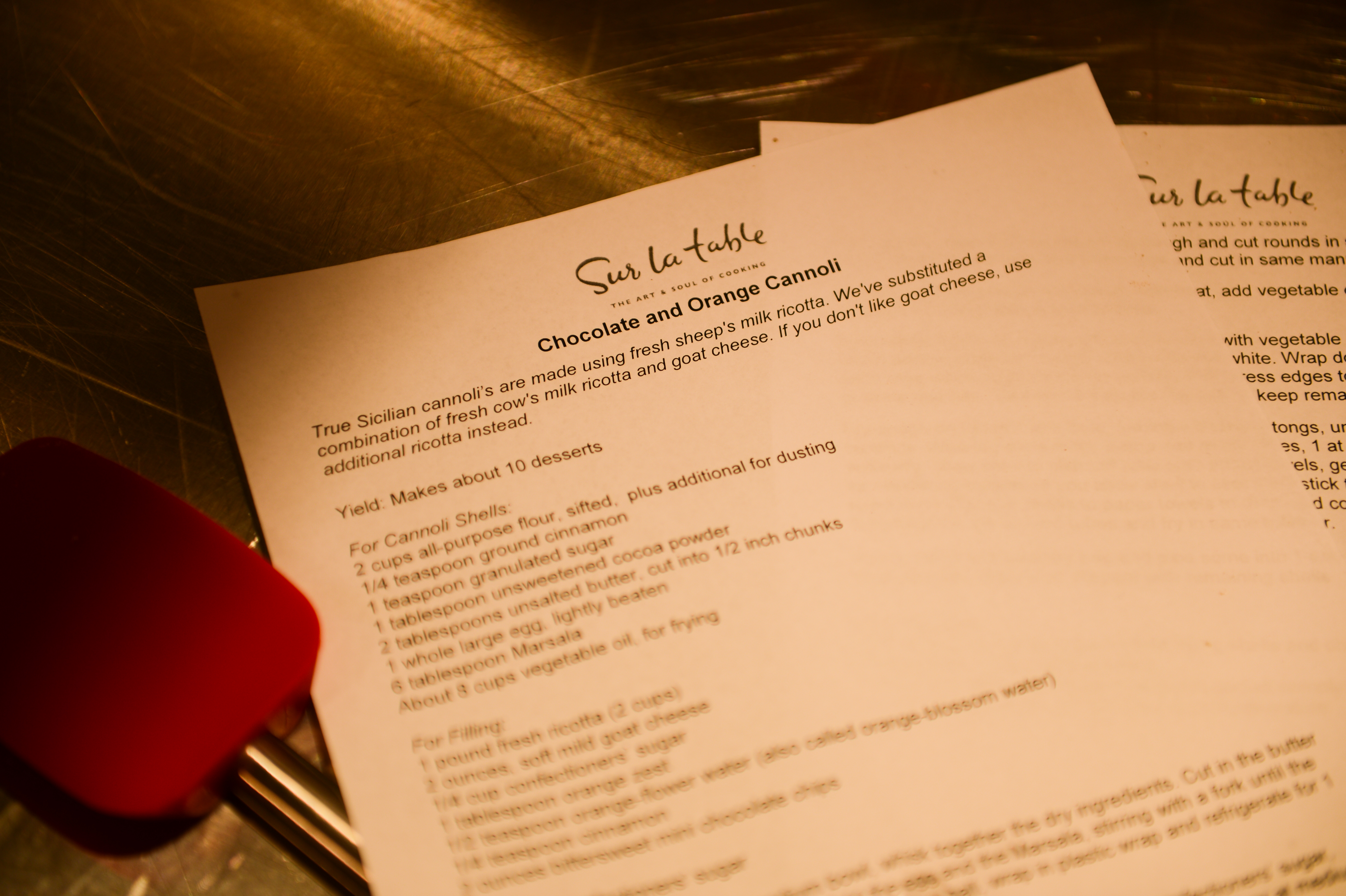 A printed recipe for chocolate and orange cannoli rests on a stainless steel surface with a red spatula nearby.