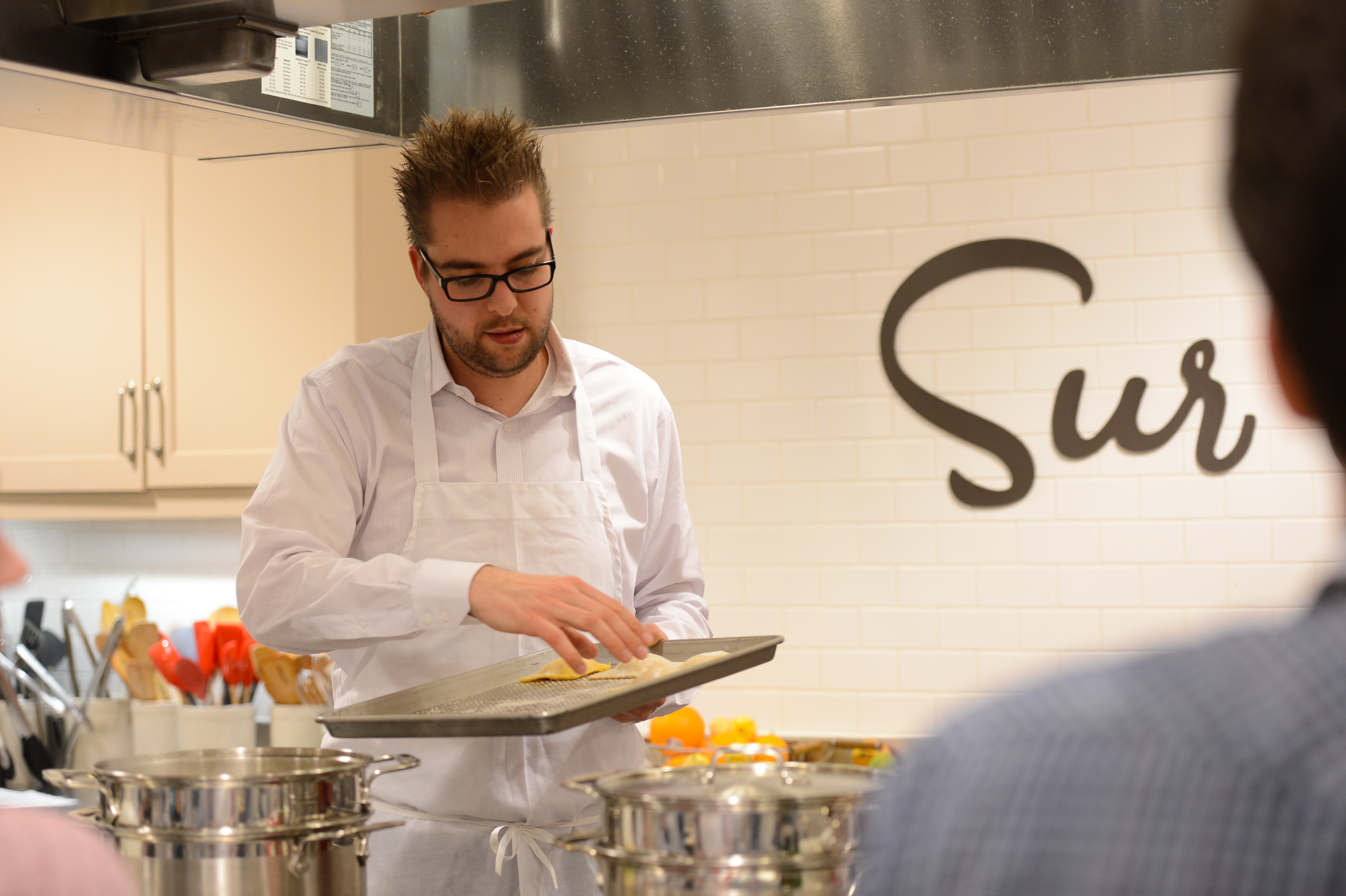 A man wearing an apron and glasses demonstrates cooking techniques while holding a tray in a kitchen classroom.