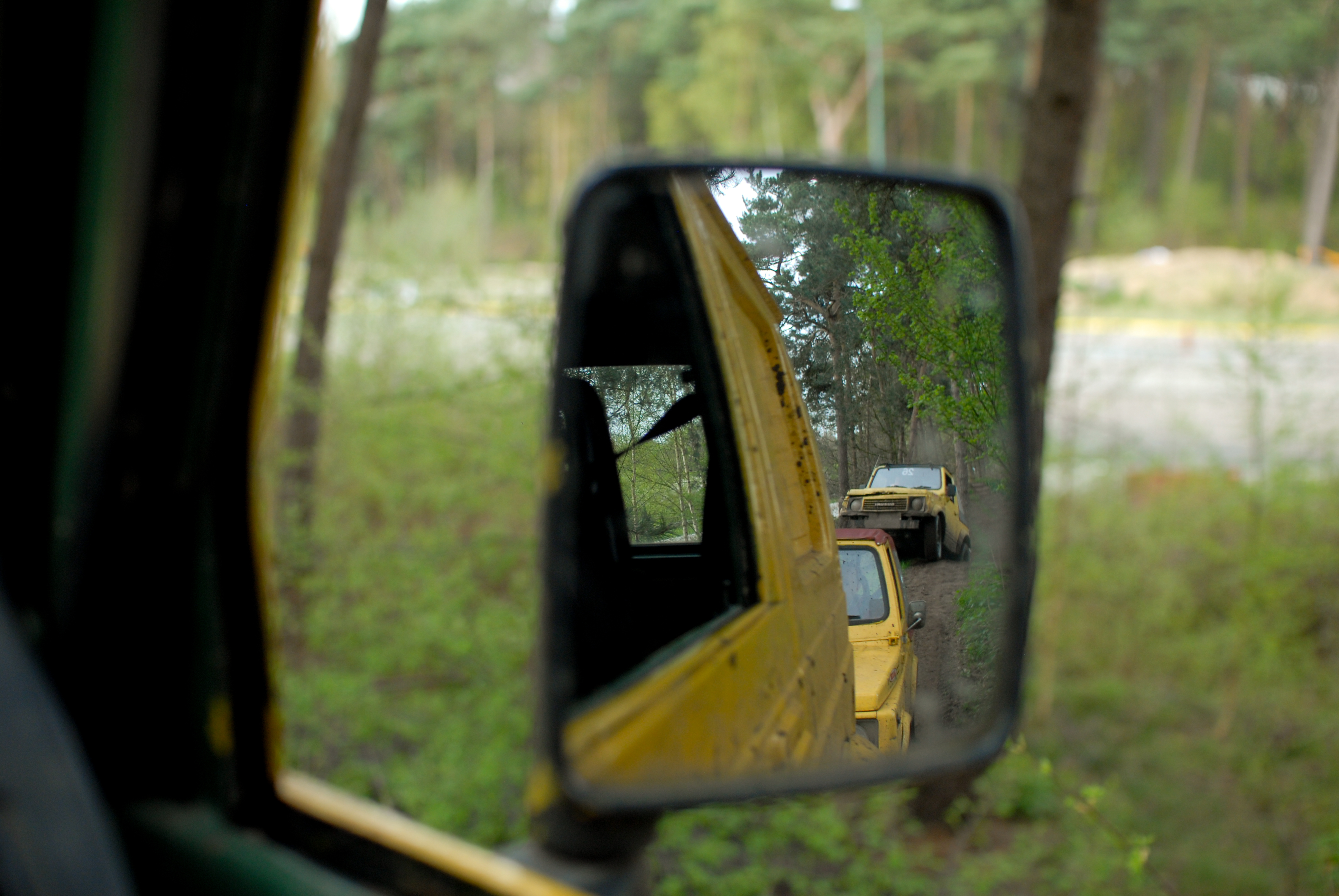 Rearview mirror reflection shows a line of yellow off-road vehicles driving on a dirt trail through the forest.