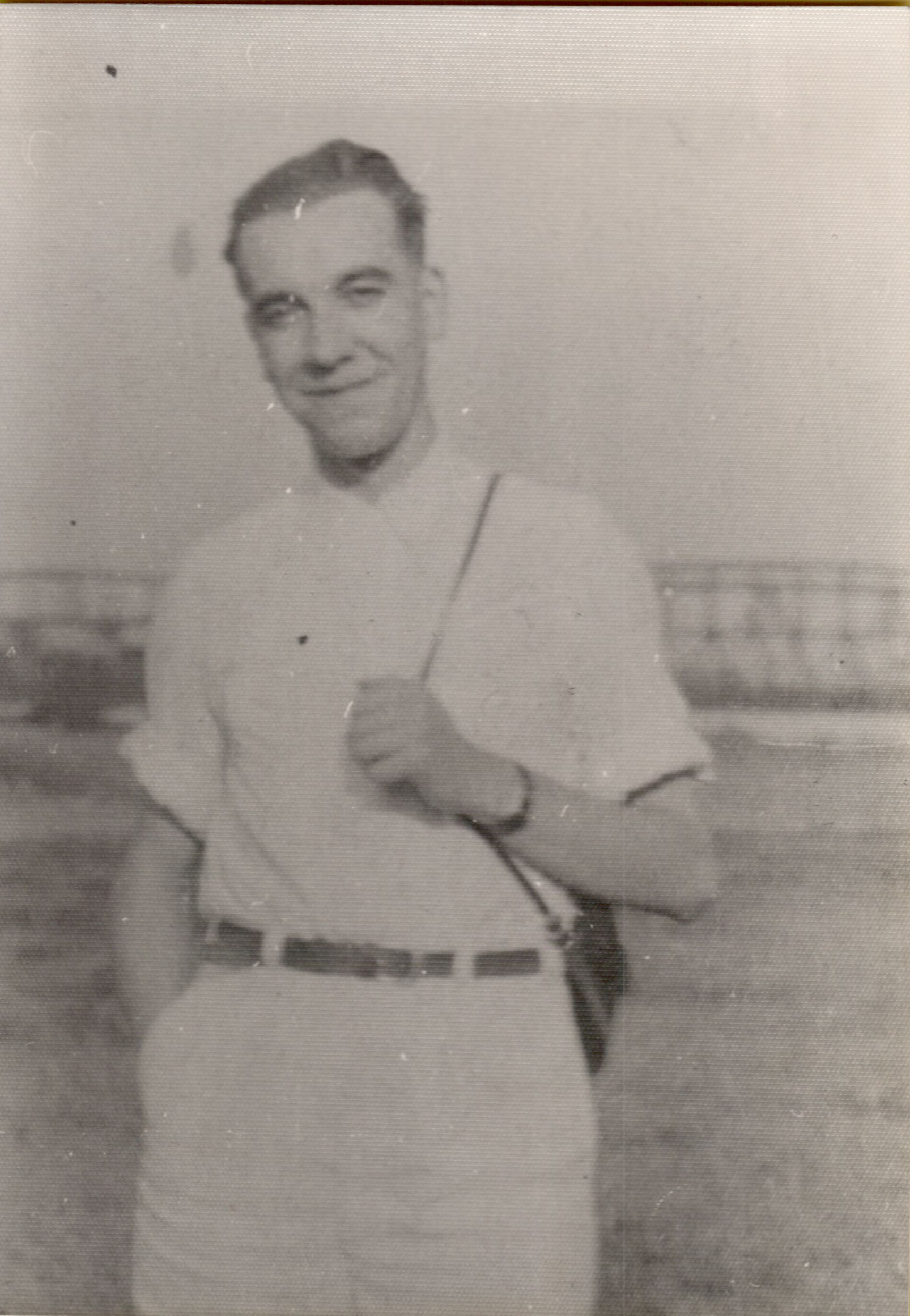 A man in a short-sleeved shirt and belt stands outdoors, smiling, with one hand in his pocket.